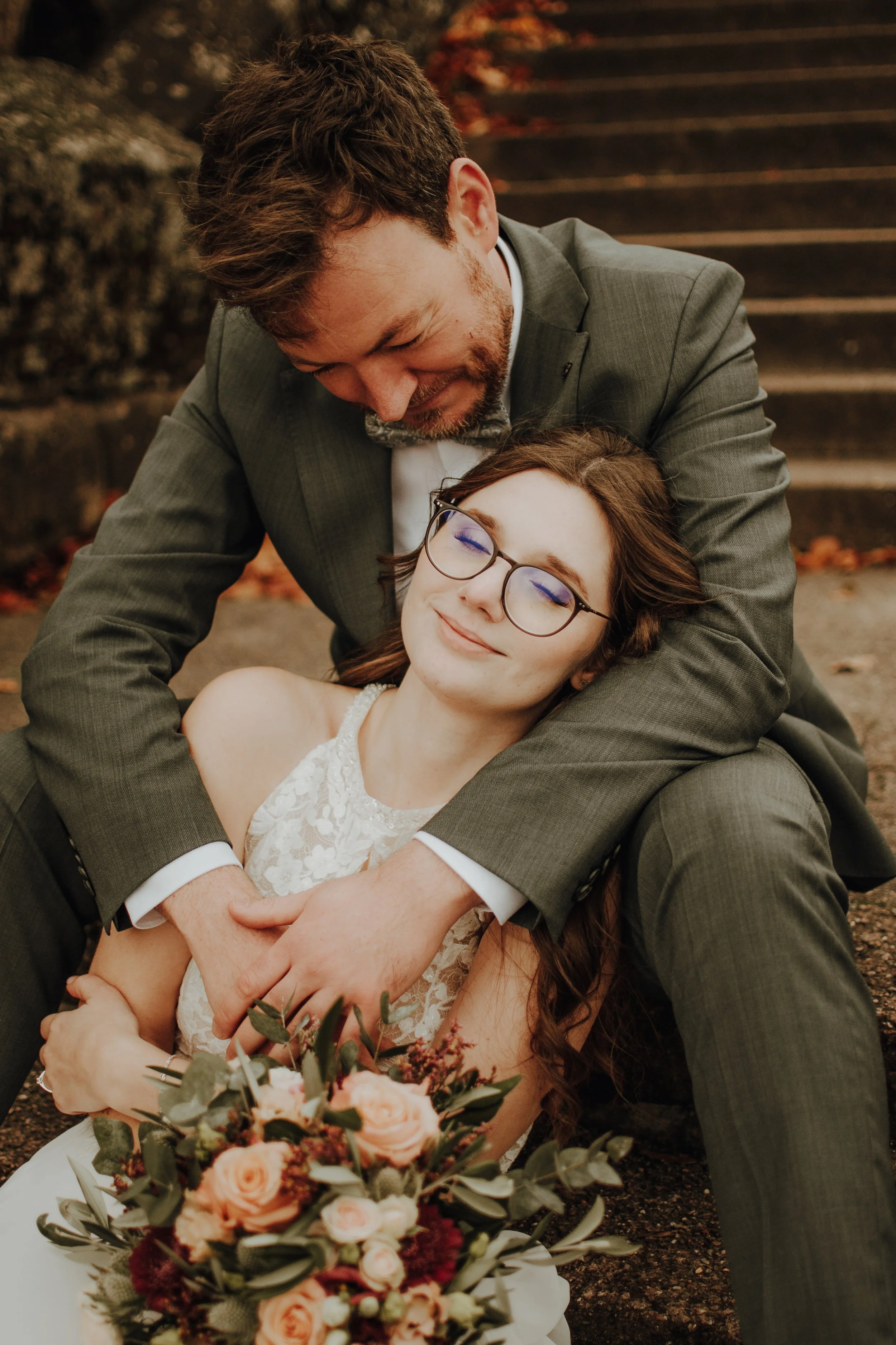 Ein Mann in Anzug umarmt eine lachende Frau in Brautkleid und Brille, während sie auf den Stufen im Freien sitzen.