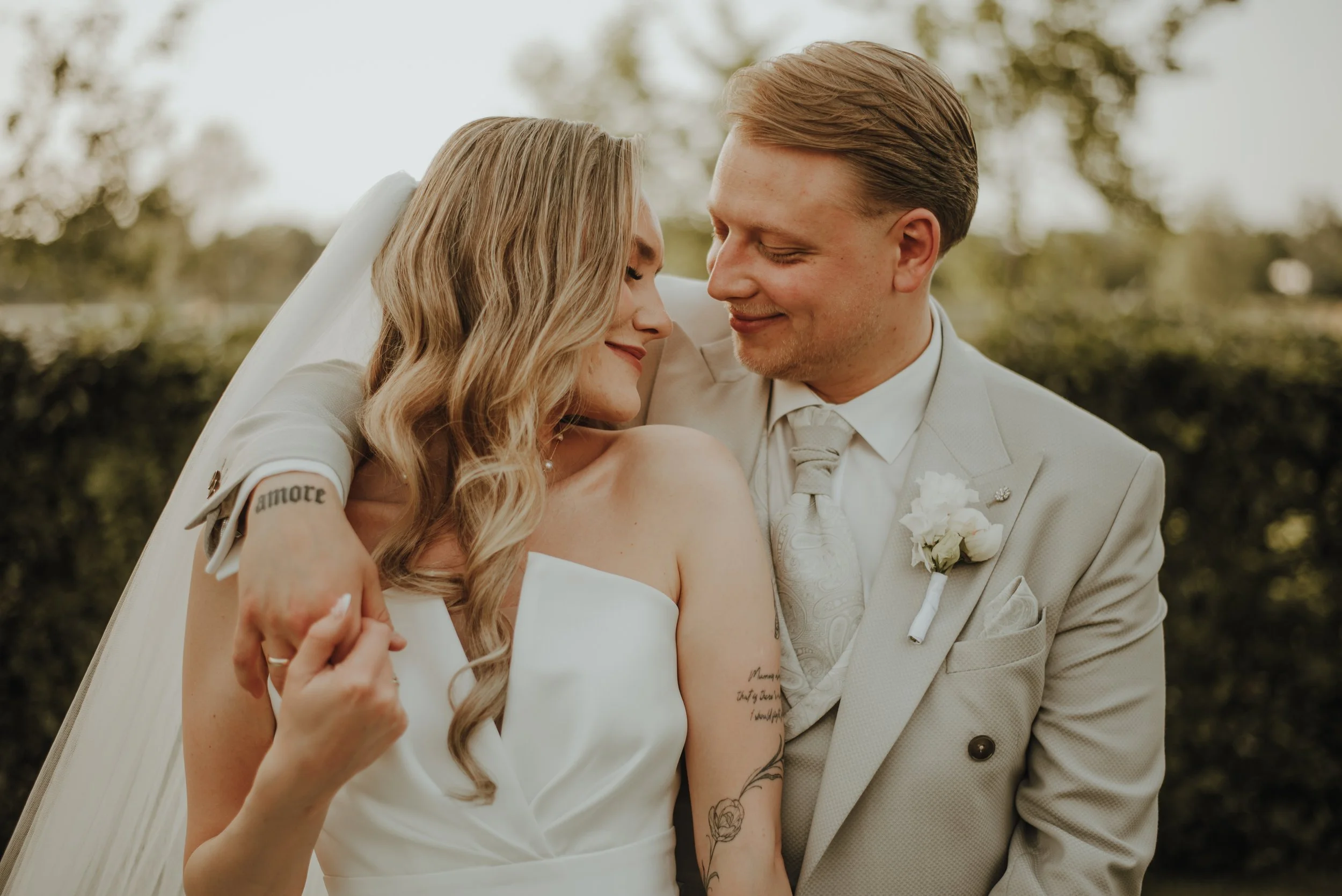 Ein Brautpaar bei einer Hochzeit, lächelnd und umarmend im Freien mit Bäumen im Hintergrund, das Brautkleid und Anzug trägt.