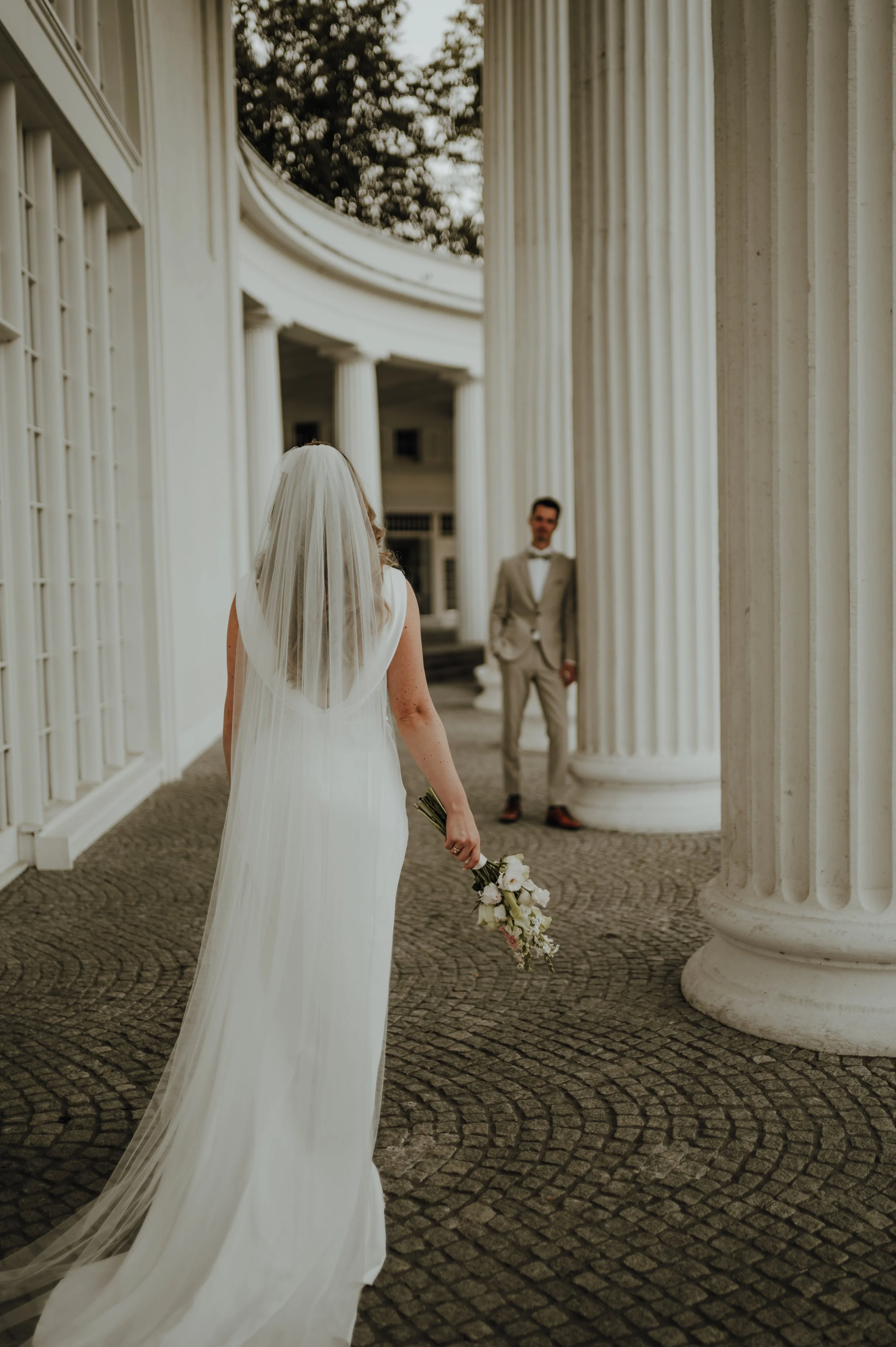 Braut in weißem Kleid mit Schleier hält Blumen, Blick auf Bräutigam vor antiken Säulen in Säulengang.