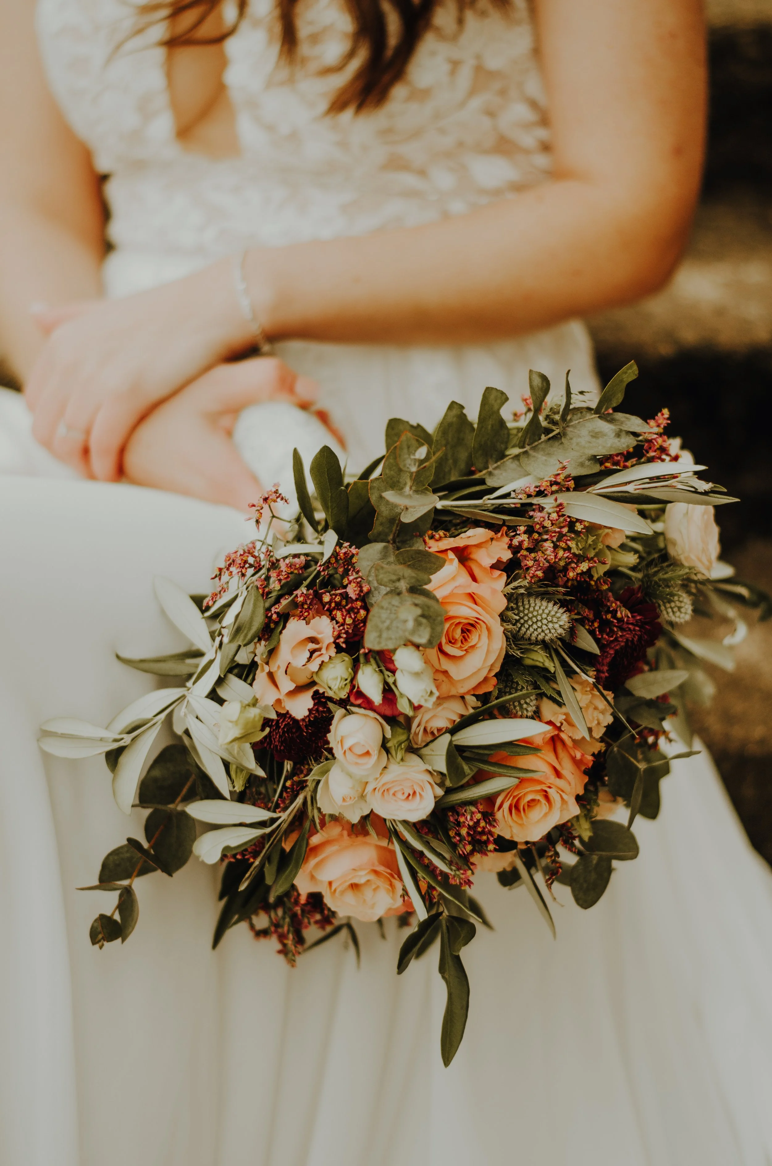 Nahaufnahme eines Brautstraußes mit orange Rosen, weißen Blumen und grünen Blättern, gehalten von einer Person in einem weißen Kleid mit Spitzenbesatz.