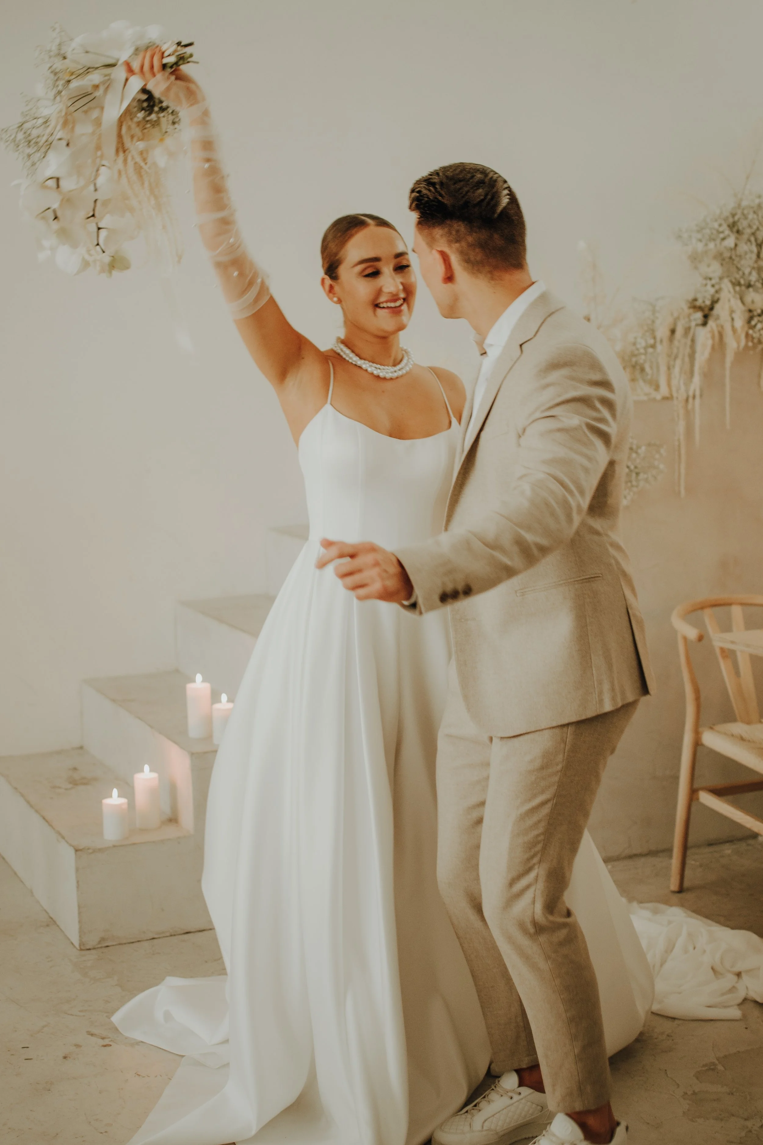 Ein Brautpaar tanzt bei einer Hochzeit, die Braut trägt ein weißes Kleid und Perlenkette, der Bräutigam einen hellen Anzug. Im Hintergrund sind weiße Kerzen auf Stufen. Die Szene ist in einem hellen, minimalistischen Raum mit weißen und neutralen Farben.