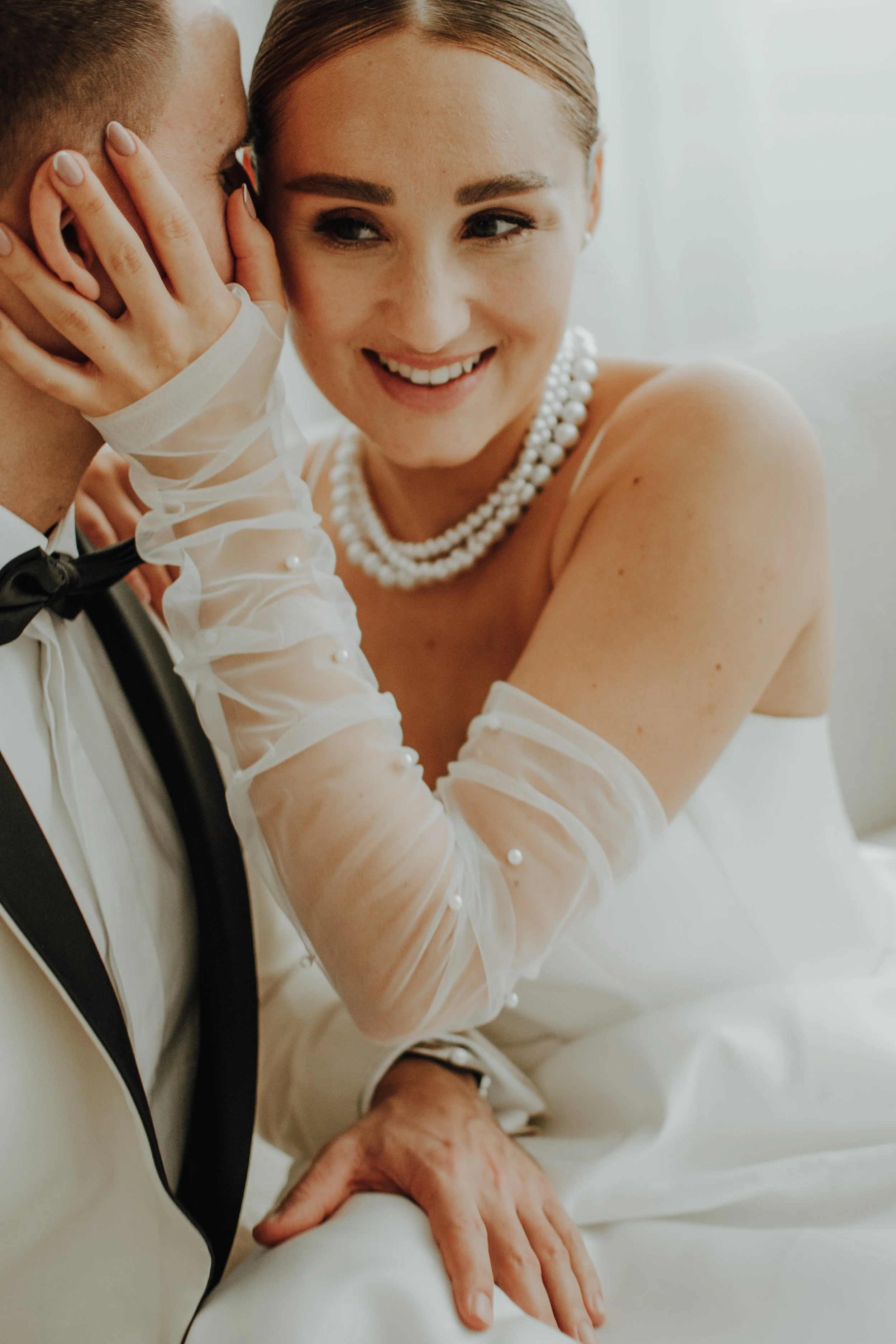 Nahaufnahme eines romantischen Moments zwischen zwei Menschen, vermutlich vor einer Hochzeit, mit einer Frau, die andeutet, dass sie vielleicht eine Braut ist, während sie eine Hand auf die Hand einer Person legt, die einen Anzug trägt. Die Frau trägt eine Perlenkette und hat ein freundliches Lächeln.