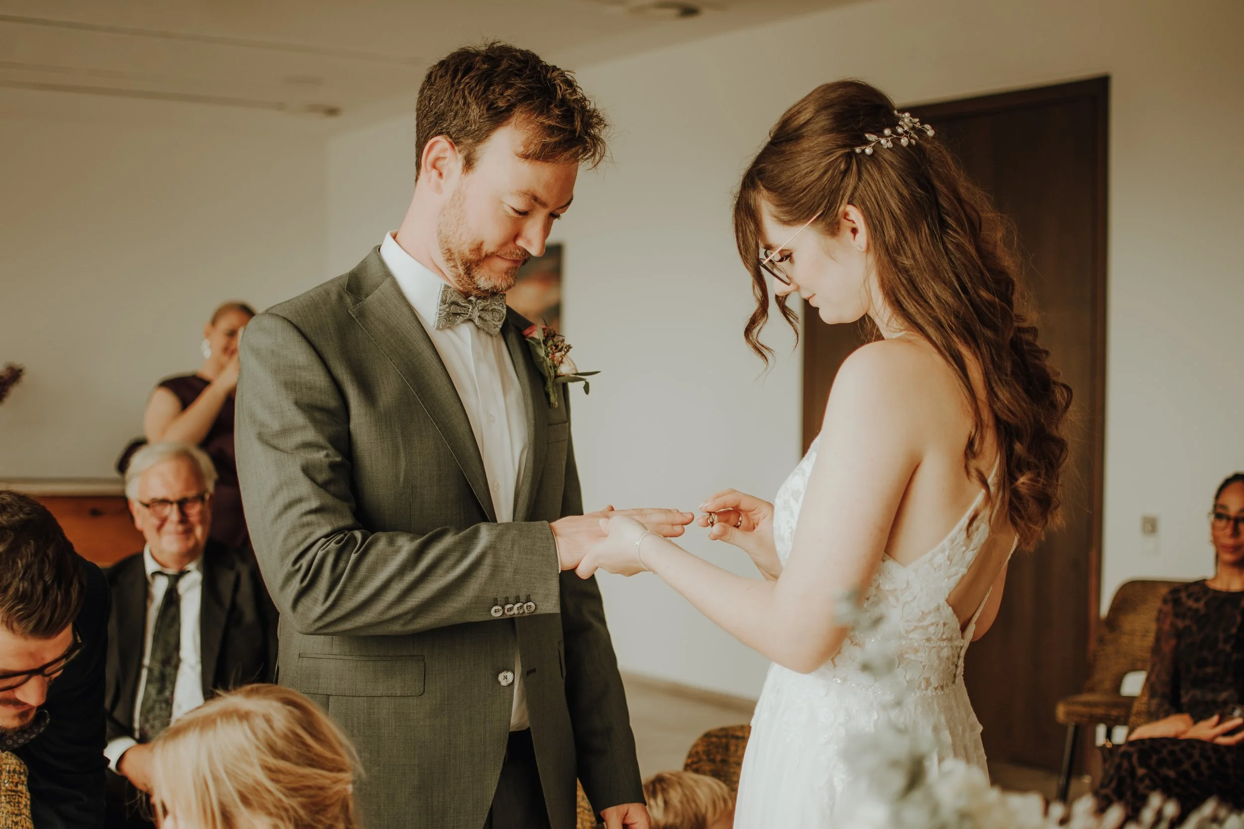 Ein Mann und eine Frau bei einer Hochzeitszeremonie, bei der sie sich die Ringe austauschen. Die Frau trägt ein weißes Brautkleid und eine dekorative Haarspange, der Mann trägt einen grauen Anzug mit Fliege.
