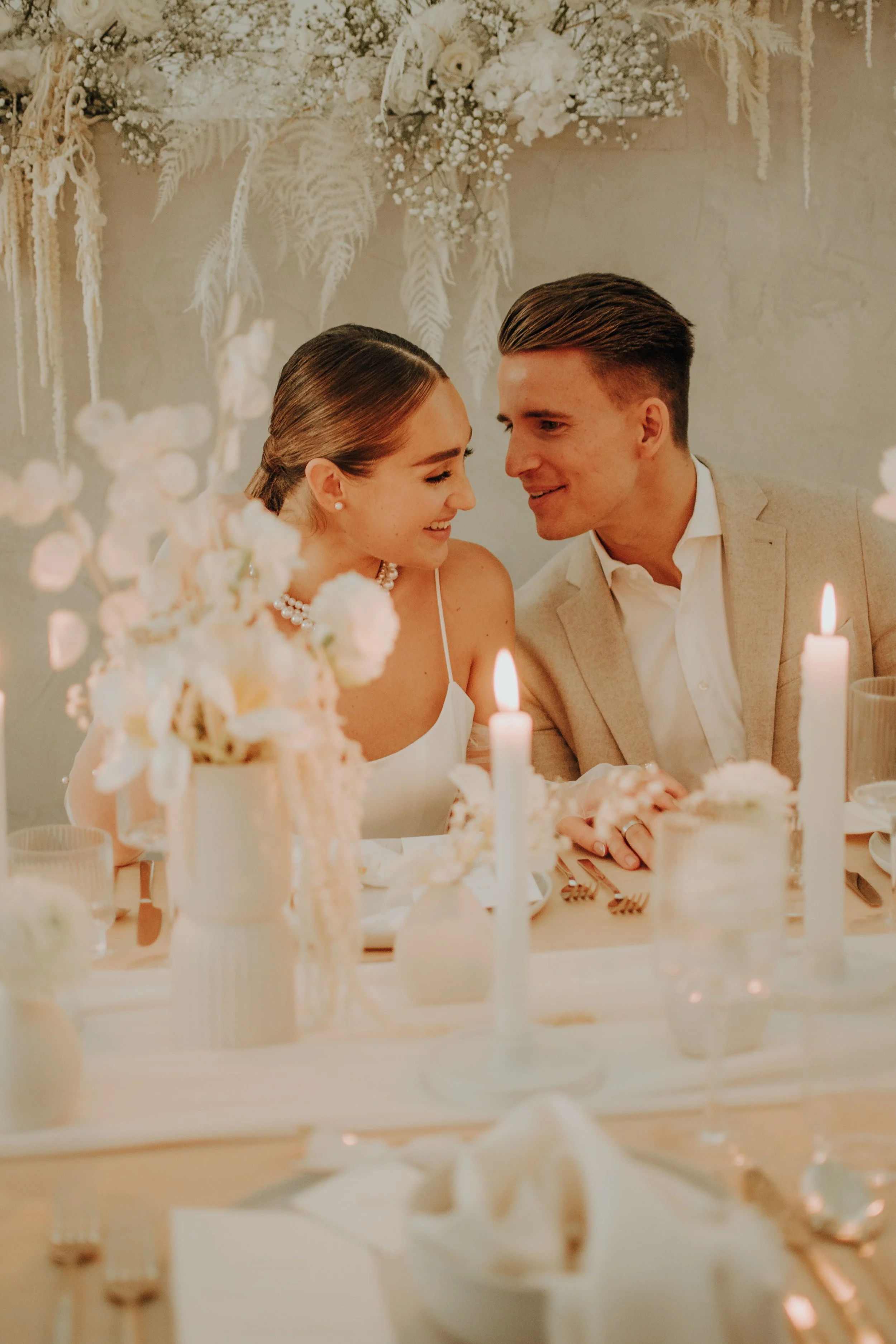 Ein Brautpaar bei ihrer Hochzeit. Sie sitzt an einem festlich gedeckten Tisch mit Kerzen und weißen Blumen. Im Hintergrund hängen weiße Blumen und Dekorationen an der Wand.