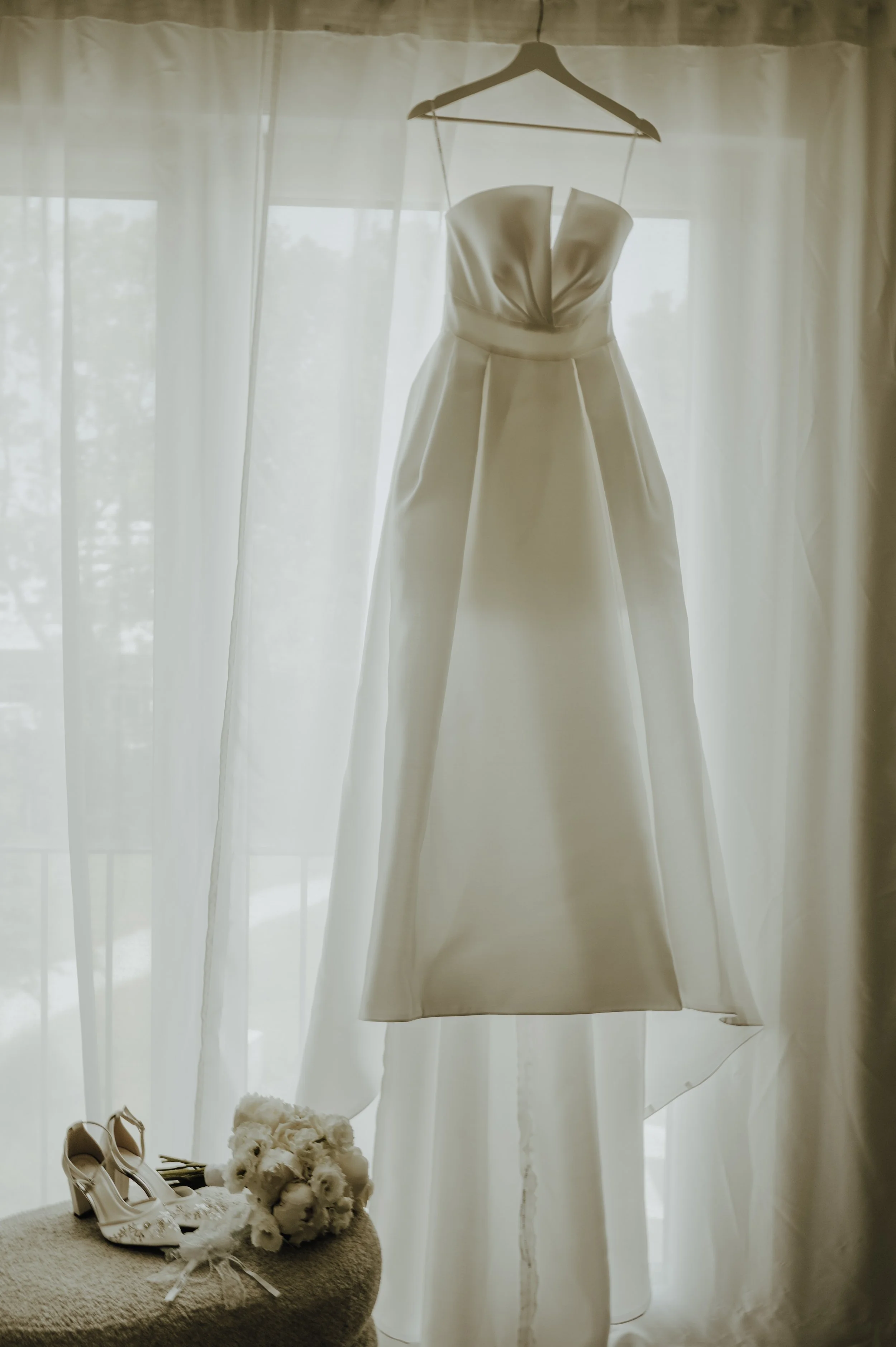 Hochzeitskleid hängt an einem Bügel vor einem Fenster mit weißen Vorhängen. Auf einem kleinen runden Tisch liegen weiße Hochzeits-High-Heels, ein Blumenstrauß und weißes Tüll. Ein weißer Teddybär liegt ebenfalls auf dem Tisch.
