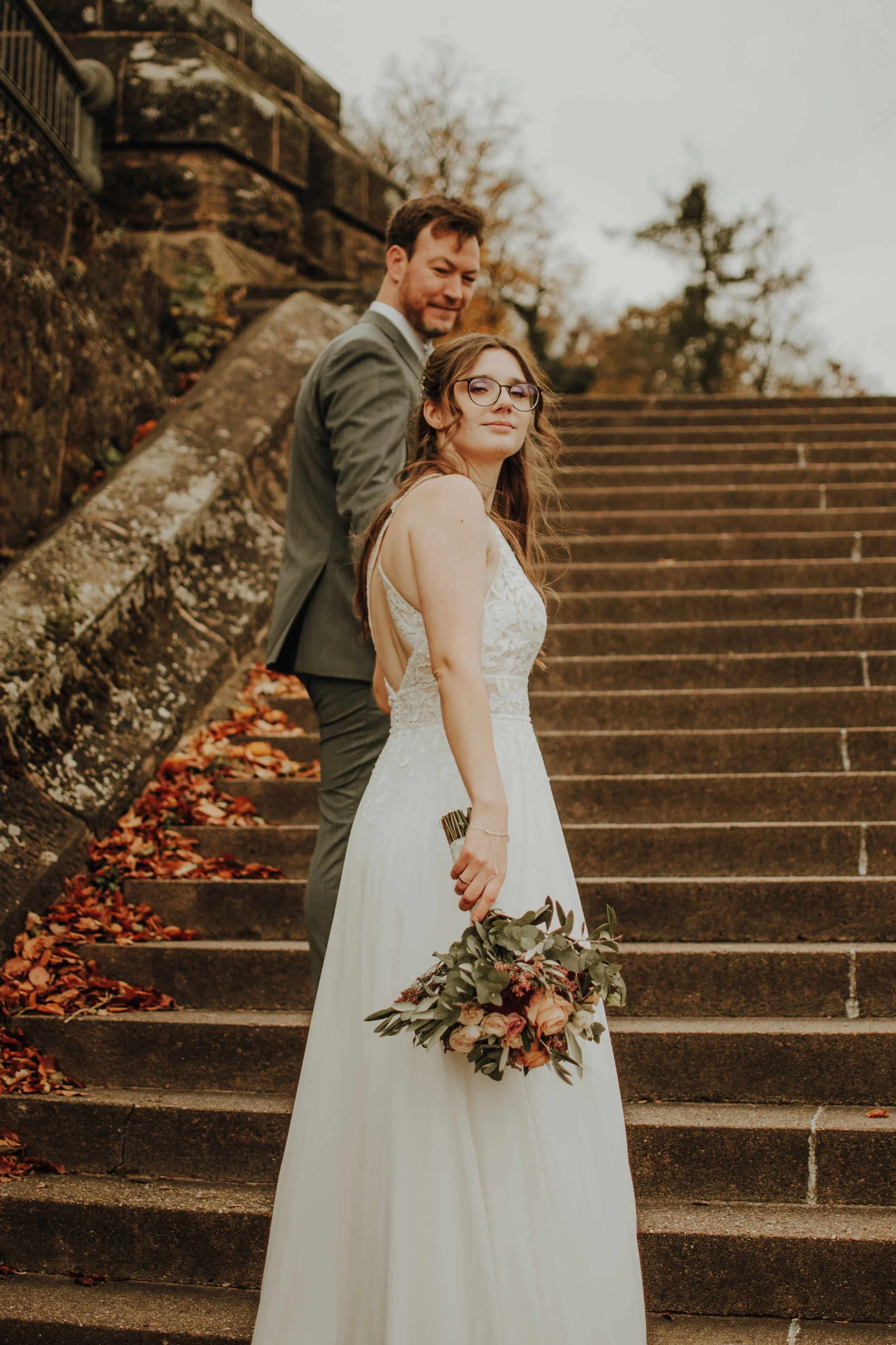 Ein Hochzeitsfotograf zeigt ein Brautpaar auf einer Treppe im Freien an einem Herbsttag, mit herabfallenden Blättern und Bäumen im Hintergrund.