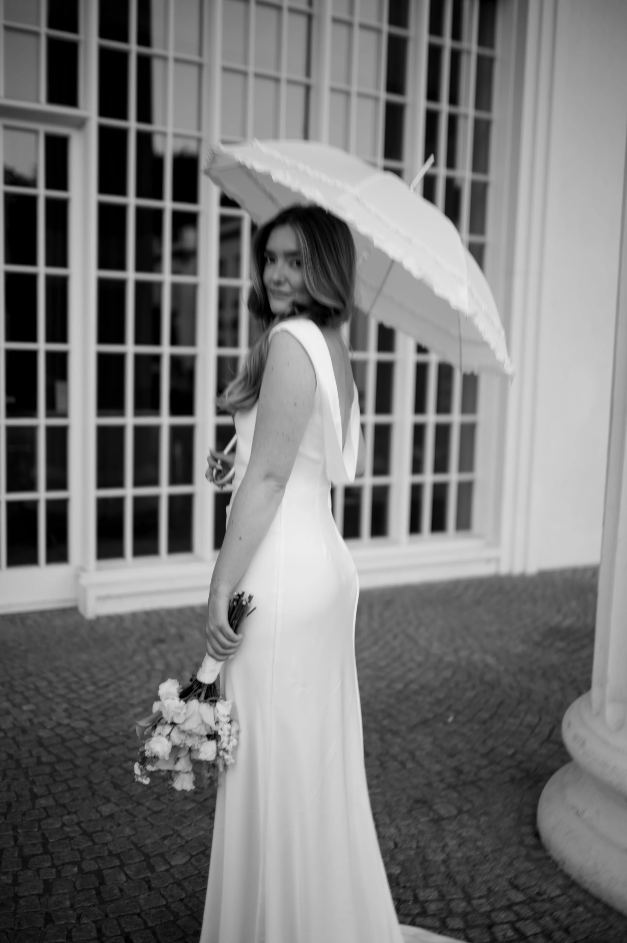 Schwarz-weiß-Foto eines jungen Frauen, die ein langes weißes Kleid, einen weißen Hut und einen weißen Regenschirm trägt, während sie vor einem Gitter steht und einen Blumenstrauß hält.