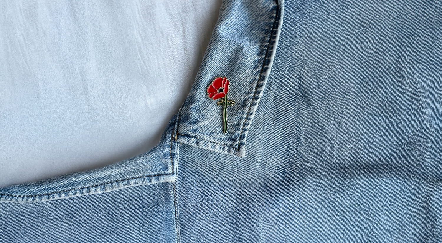 A close-up of a denim jacket with a red Crown Anemone pin attached near the collar.