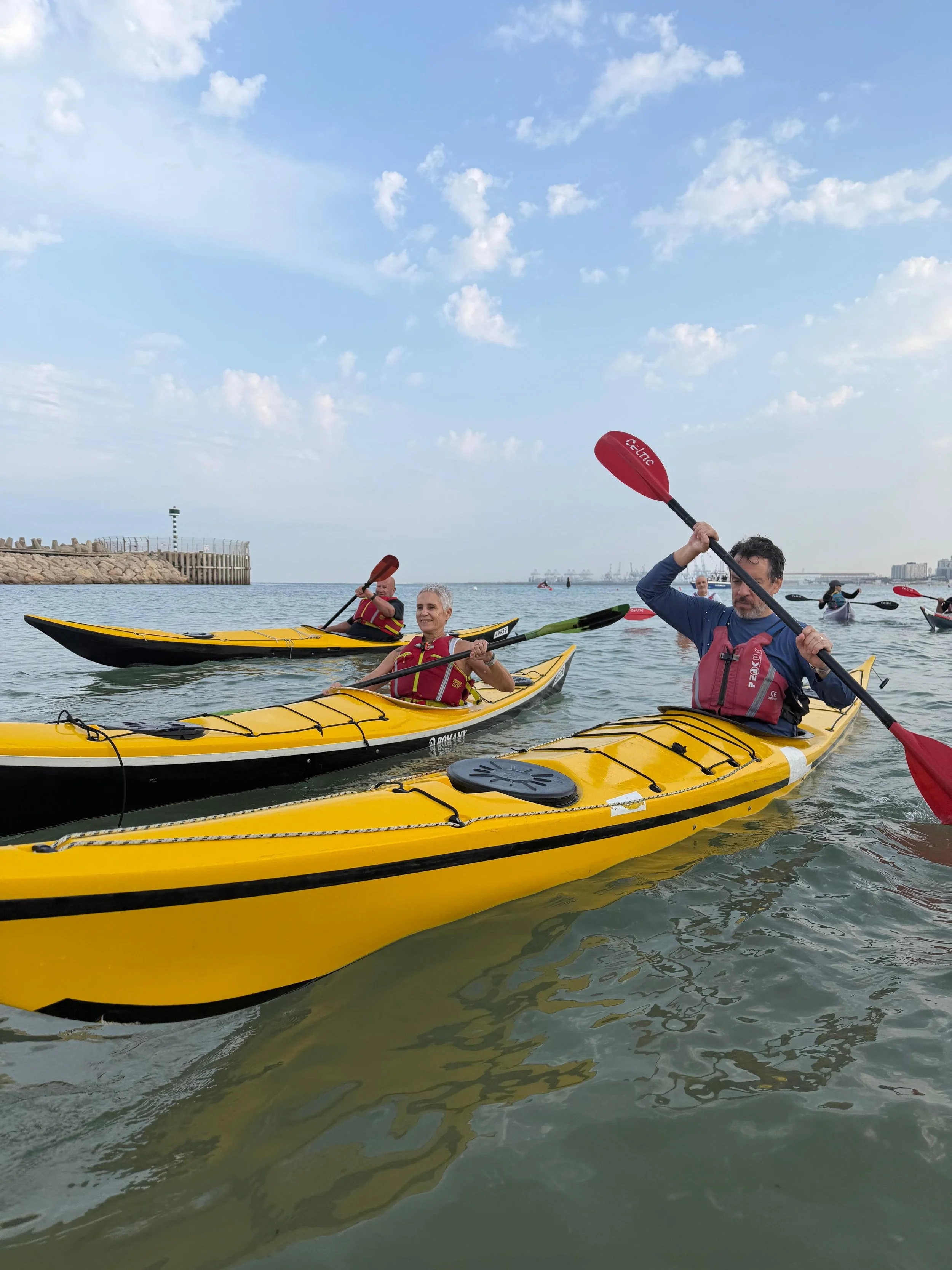 Group of people kayaking on water with a cityscape and a lighthouse in the background under a partly cloudy sky.