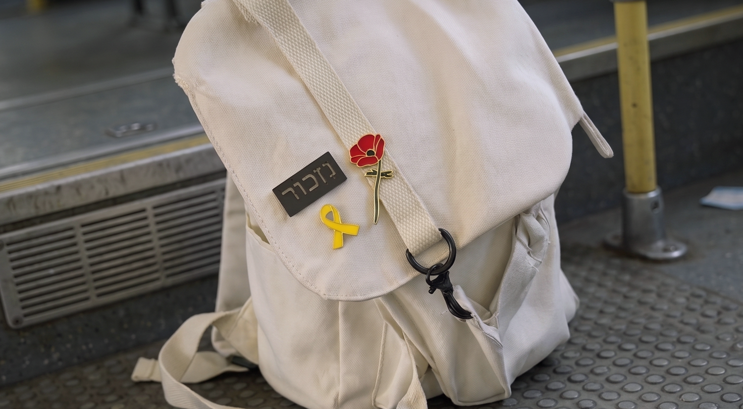 A white backpack with three pins attached: a black rectangular pin with Hebrew letters, a yellow ribbon pin, and a red Crown Anemone flower pin. The backpack is on the floor of a local israeli bus 