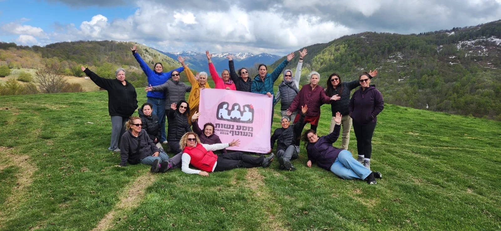 Group of women outdoors holding a pink banner with Hebrew text and graphic silhouette of women, with mountains and cloudy sky in the background.
