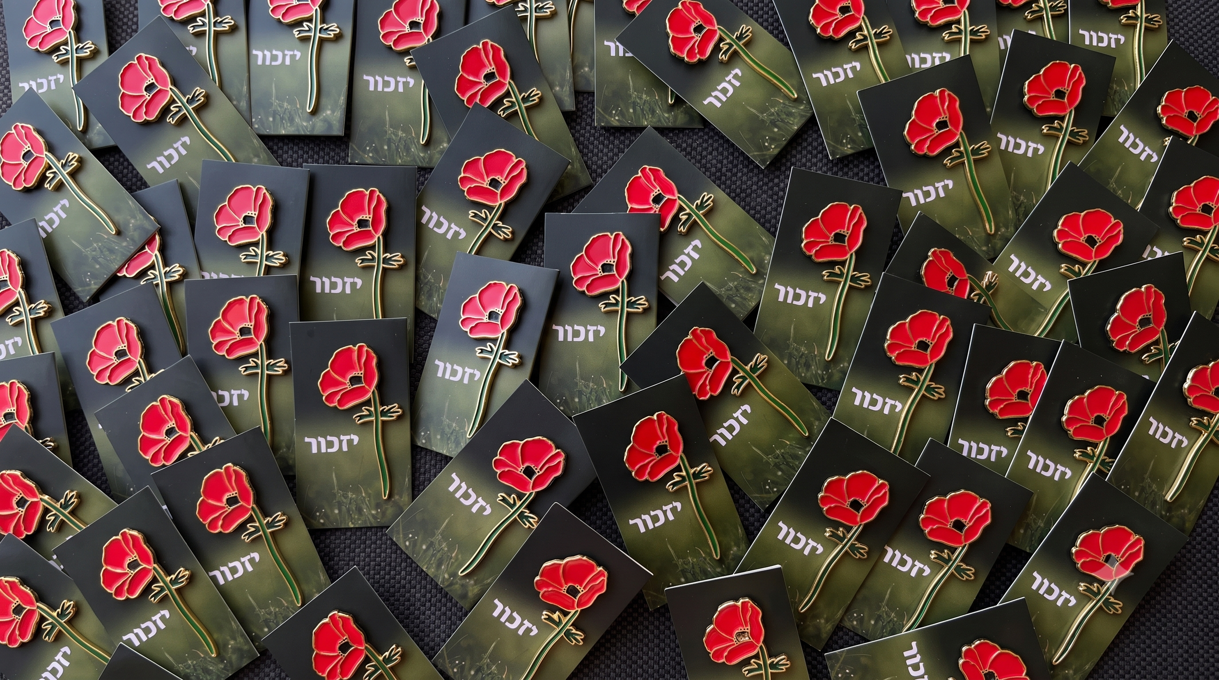 A collection of pins with red Crown Anemone flowers and Hebrew text on black and green backgrounds, scattered across a dark surface.