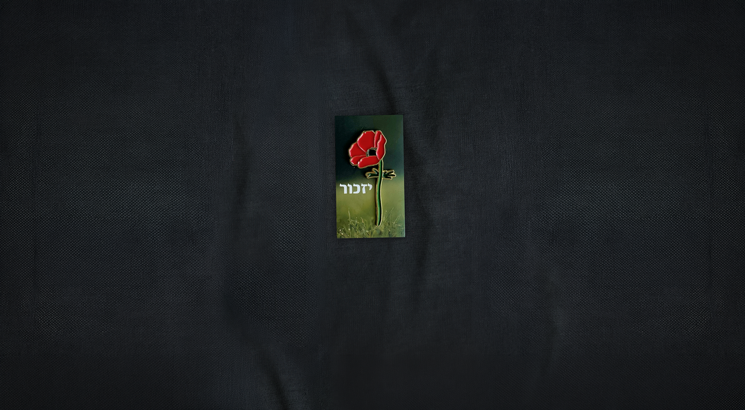 A black fabric surface with a small rectangular card featuring a red Crown Anemone flower with green stem and Hebrew text.