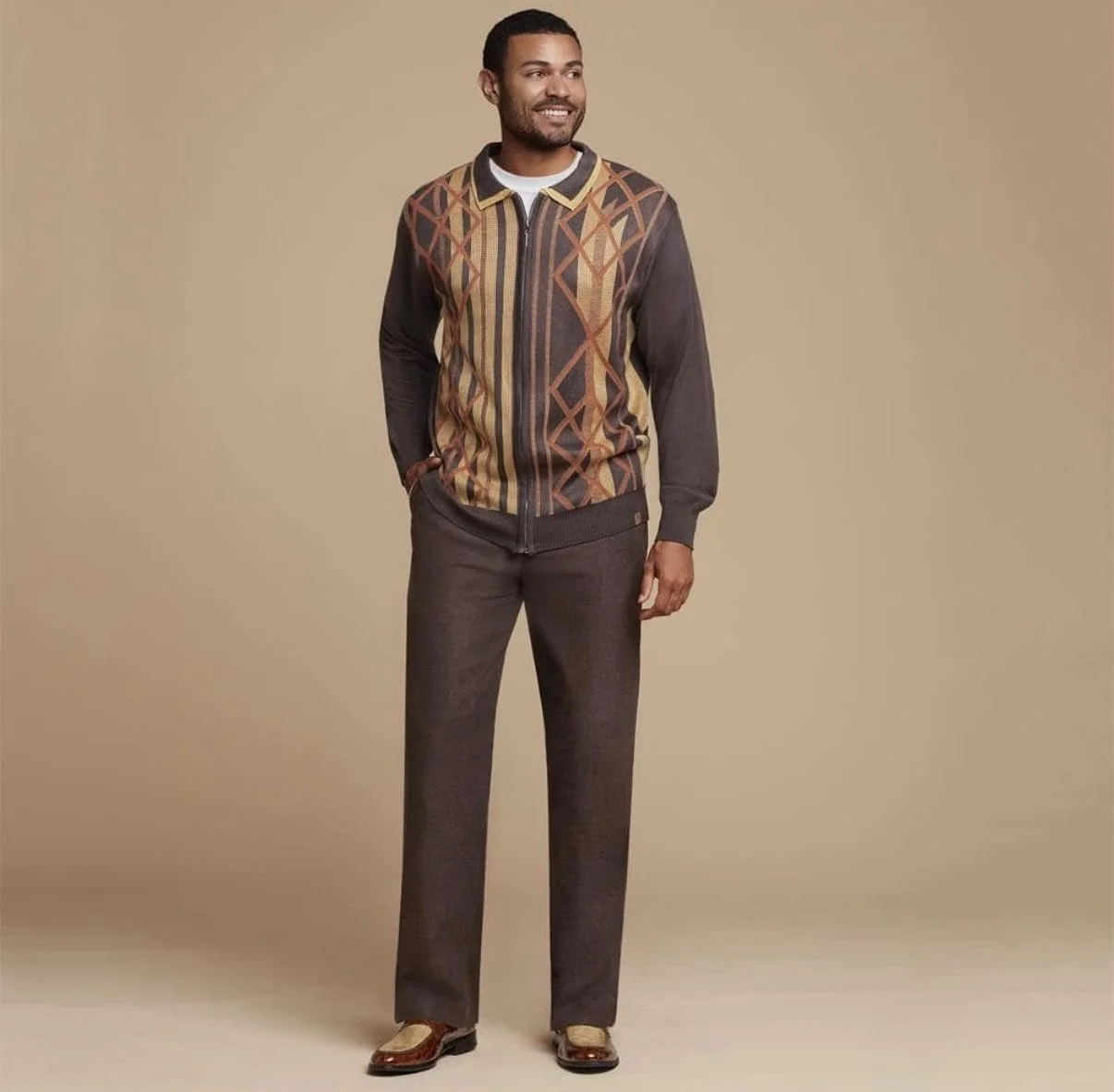 A man wearing a patterned zip-up jacket, dark pants, and brown shoes, standing against a plain beige background.