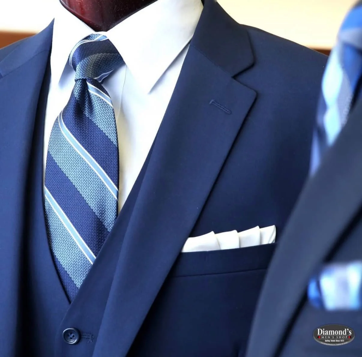 Close-up of a navy blue suit jacket, white dress shirt, and a blue striped tie with a pocket square in the chest pocket, on a mannequin at a men's clothing store.