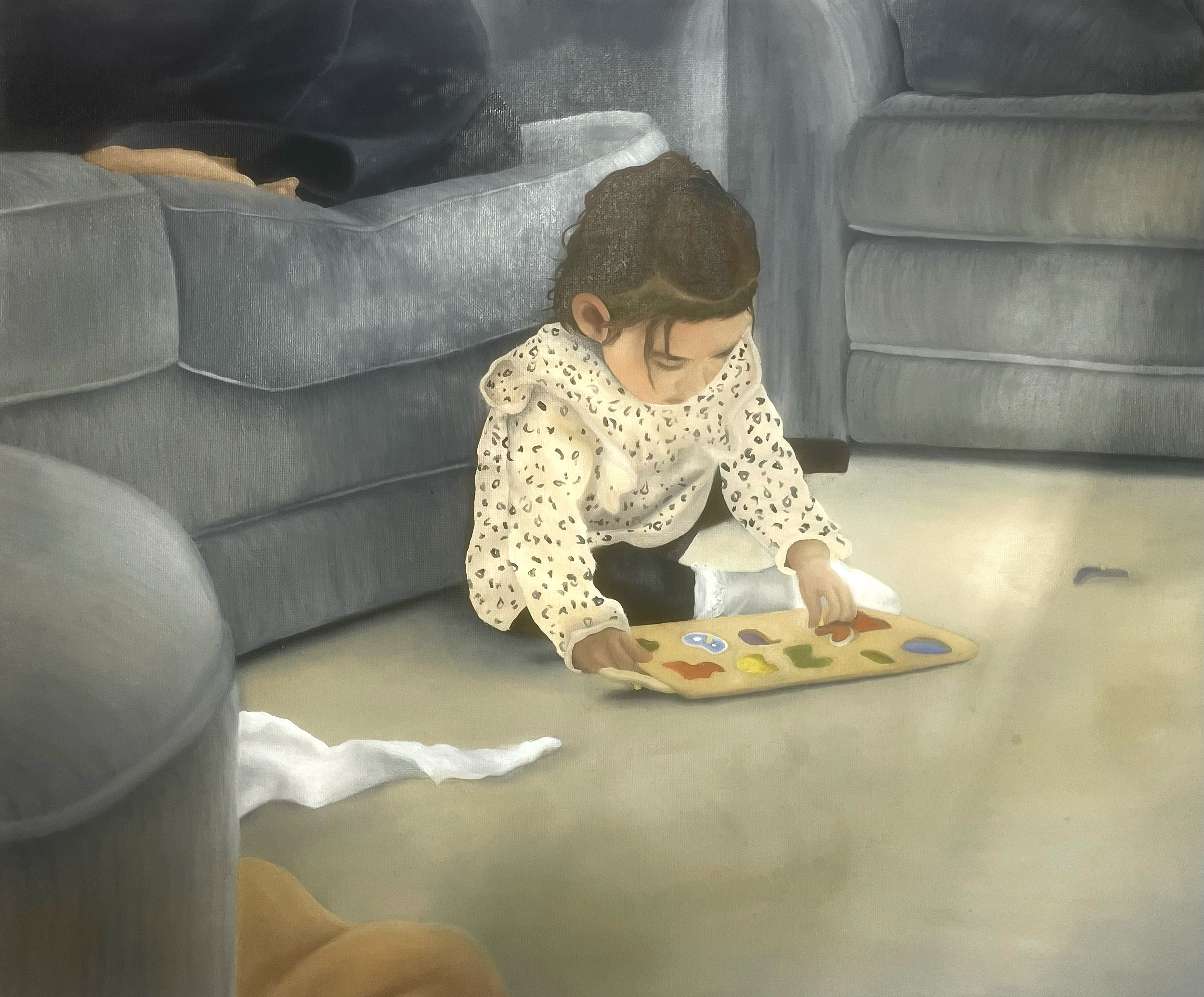 A young girl with curly hair, wearing a white polka-dot shirt, is sitting on the floor playing with a colorful puzzle board. A gray sofa is behind her in a room with light-colored flooring.