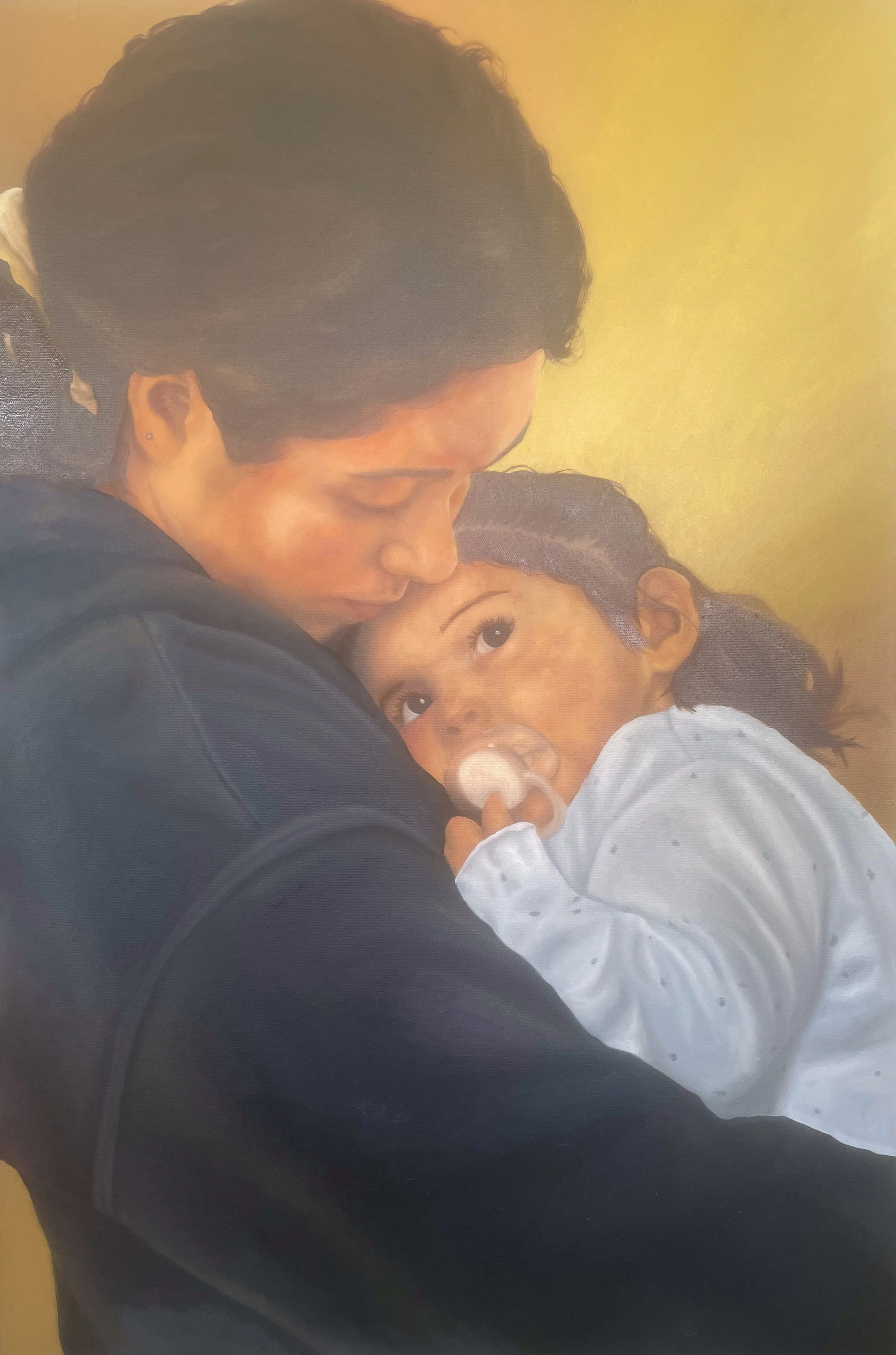 A woman and a young girl are embracing, with the girl looking up at the woman. The woman has short dark hair, a dark jacket, and small earrings. The girl has dark curly hair, a white outfit, and a pacifier in her mouth. They are against a plain yello