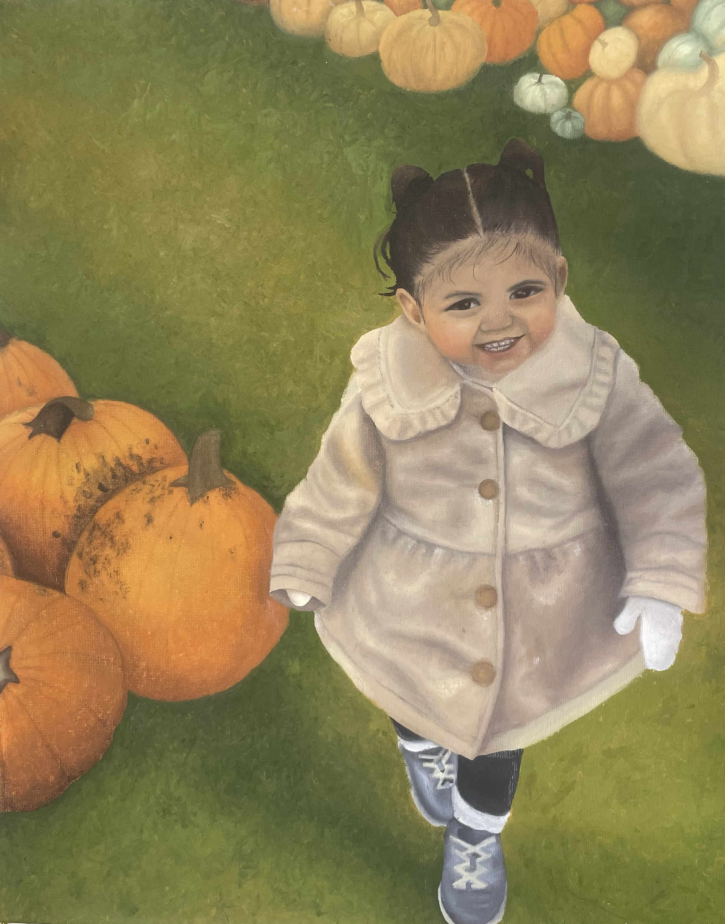 A young child with dark hair in pigtails, smiling, crouching on green grass surrounded by pumpkins, with more pumpkins in the background.