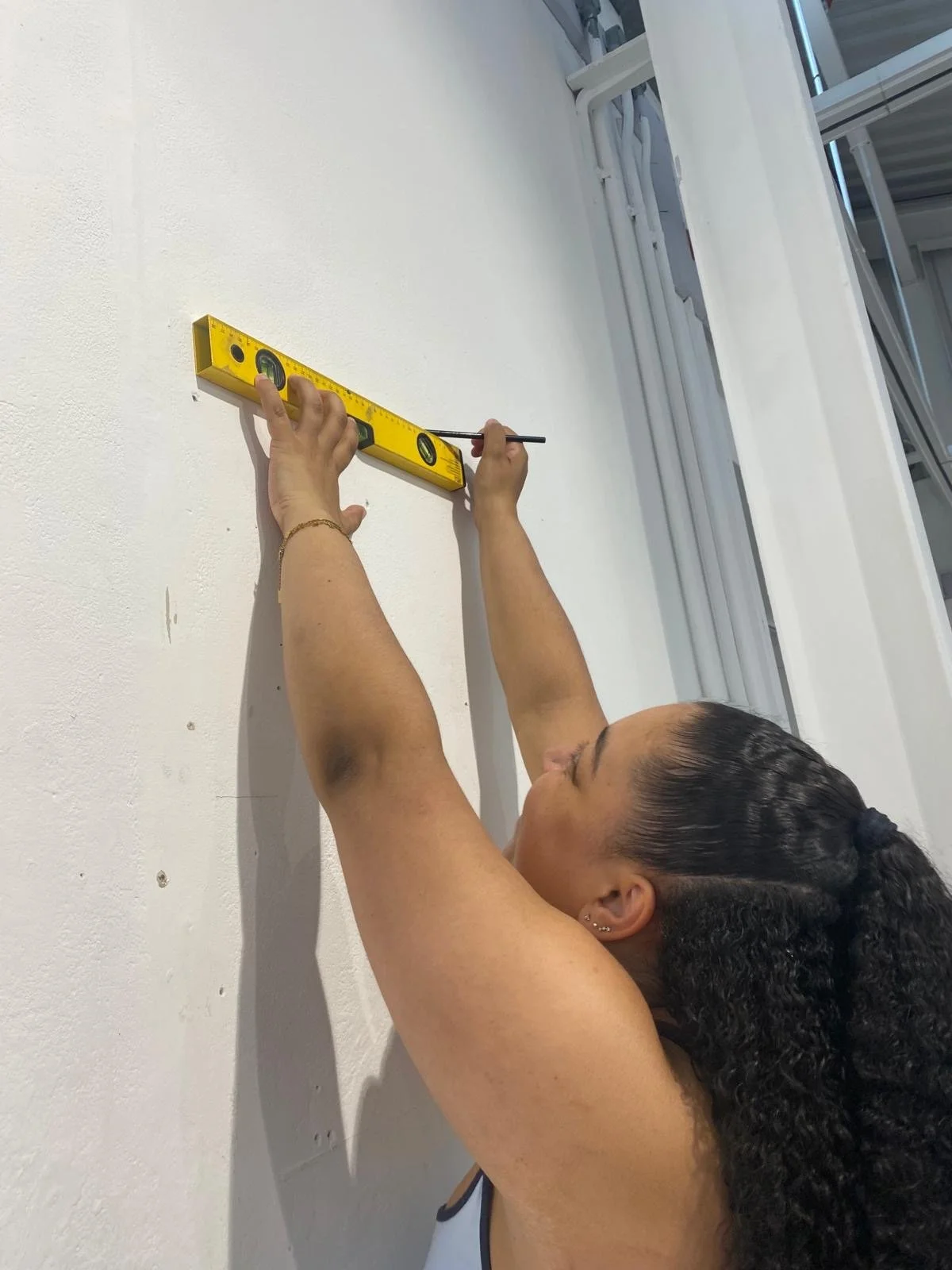 A woman with curly dark hair using a level and a small black stick to measure or mark a wall.
