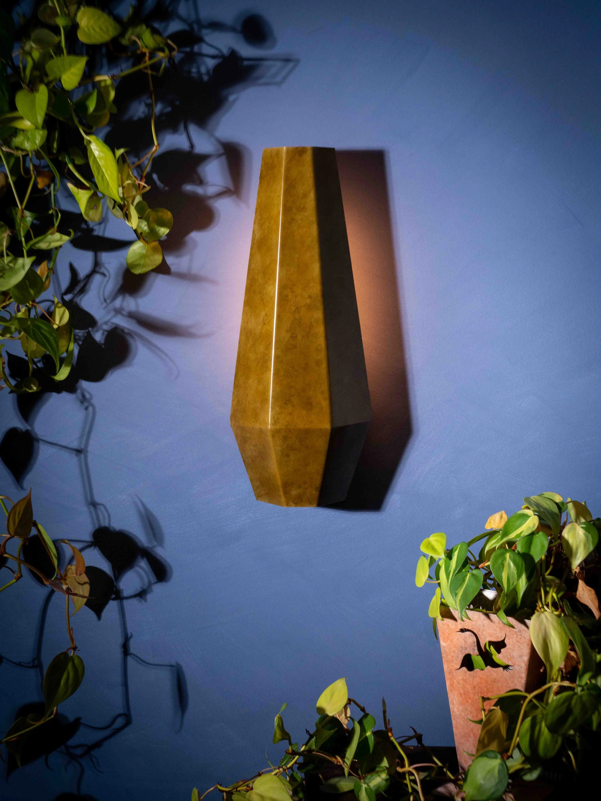 Decorative wall with a tall, narrow, gold-colored object, possibly a sculpture or speaker, surrounded by green leafy plants with shadows cast on a blue wall.