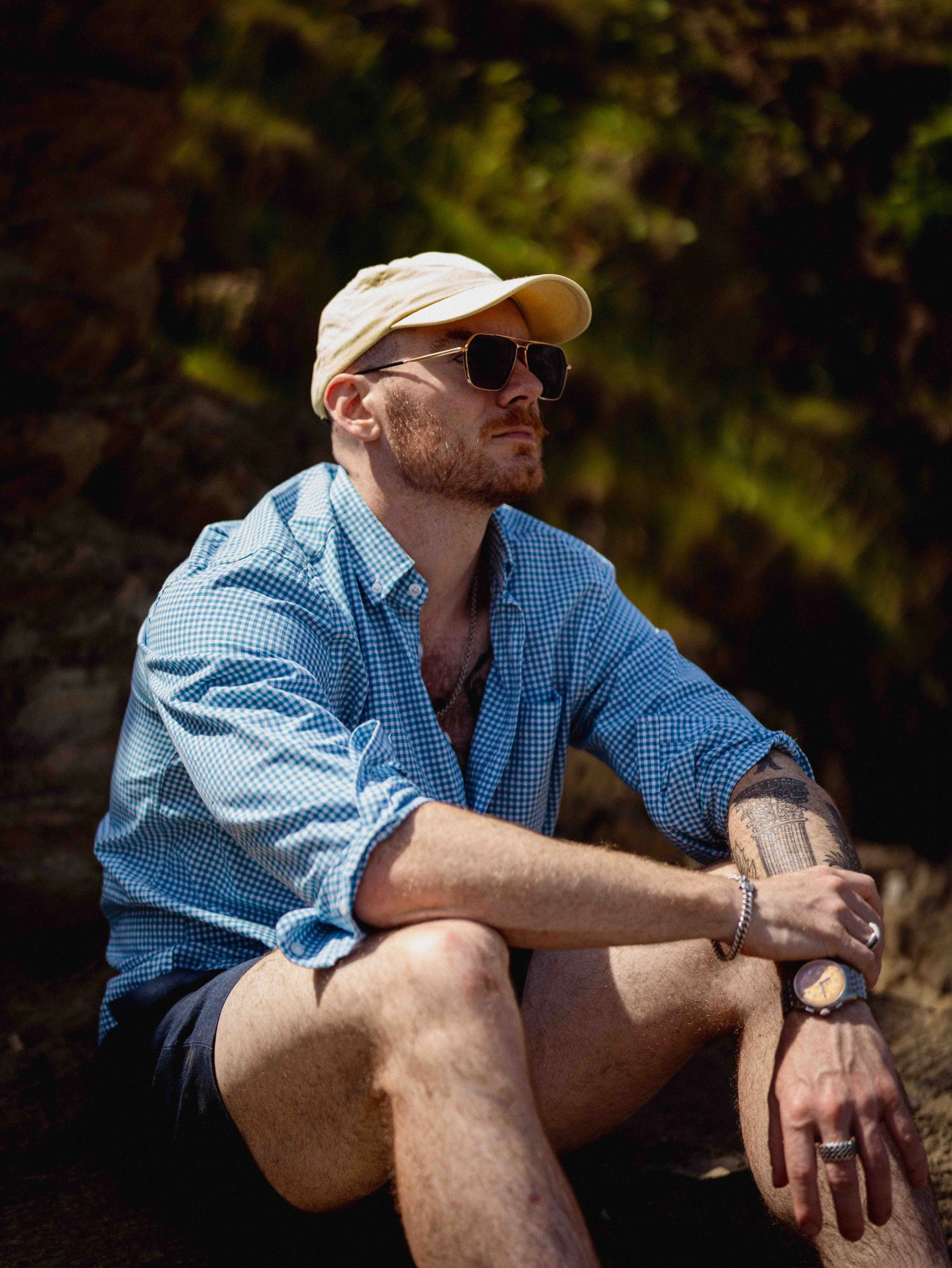 A man wearing a beige cap and black sunglasses sitting outdoors on rocks, with a tattoo on his upper arm, wearing a blue checkered shirt and a silver bracelet, during daytime.