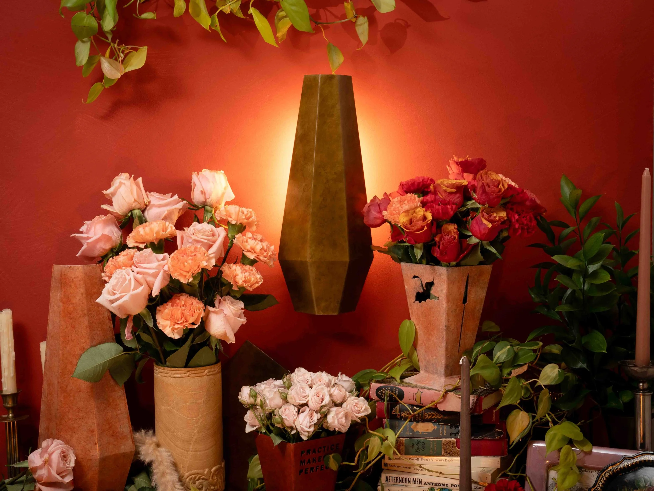 A decorative interior display features tall, gold-colored vases filled with pink and peach roses, set against a warm reddish wall with a light source behind a central gold vase. There are green leafy plants and books on a shelf below the floral arran