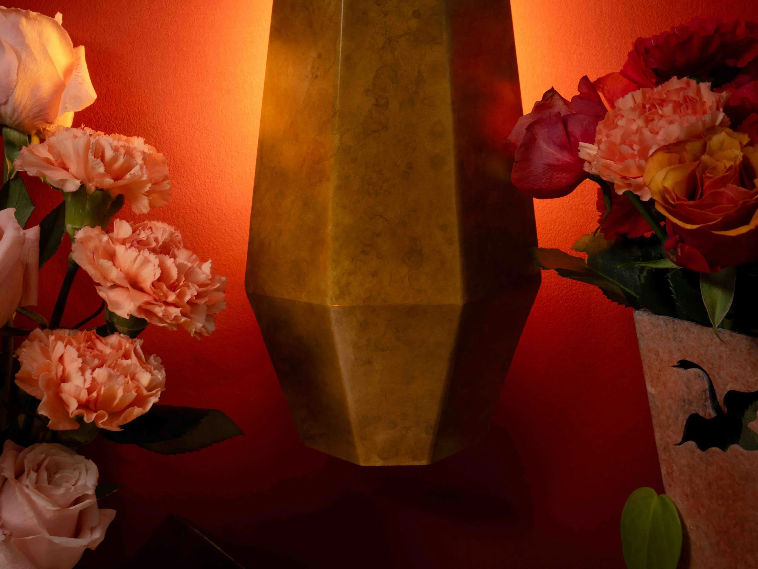 A tall, yellowish-green ceramic vase with a geometric shape, placed against an orange wall, surrounded by pink and red artificial or real flowers.