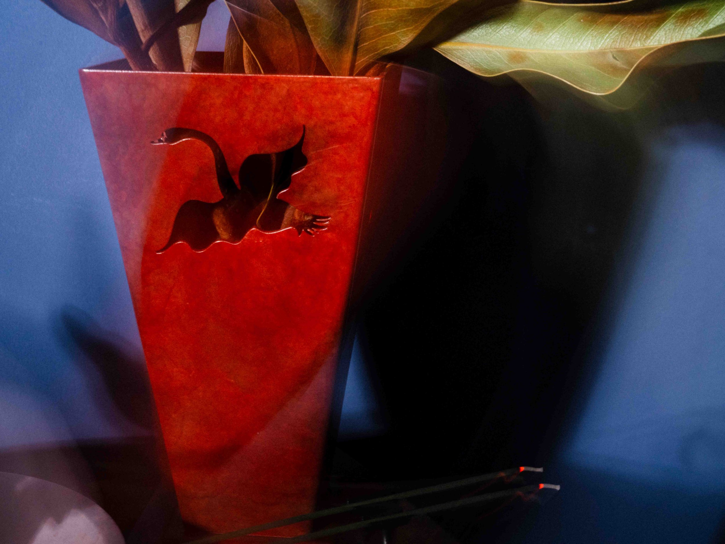 A red-shaped vase with a cutout silhouette of a woman with flowing hair, holding a flower, placed against a blue wall. Green leaves are visible behind the vase.
