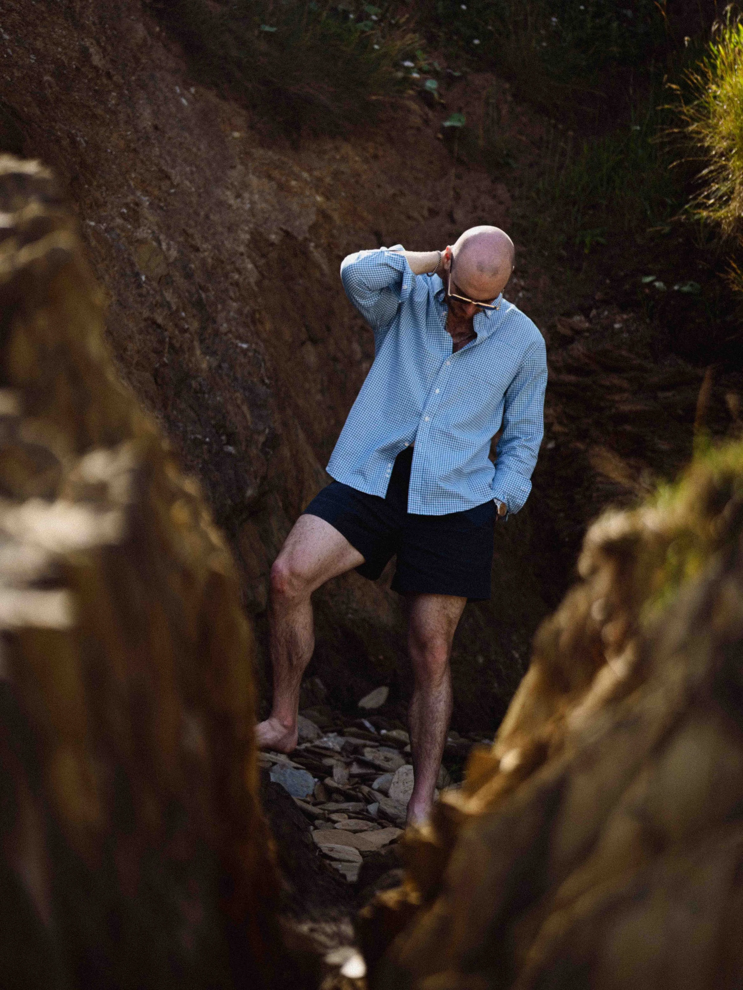 A man with a shaved head, glasses, and a beard stands outdoors in rugged terrain. He is wearing a blue checkered shirt and black shorts, and is scratching his head with his right hand. The photo is taken through a hole or opening, with the edges cove