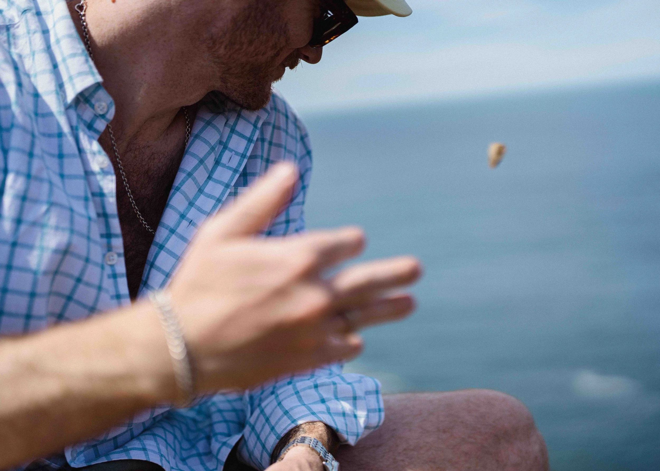 A man in a plaid shirt, wearing sunglasses, a silver chain, a beige cap, and a watch, is outdoors near water, holding a small object or insect in his hand.
