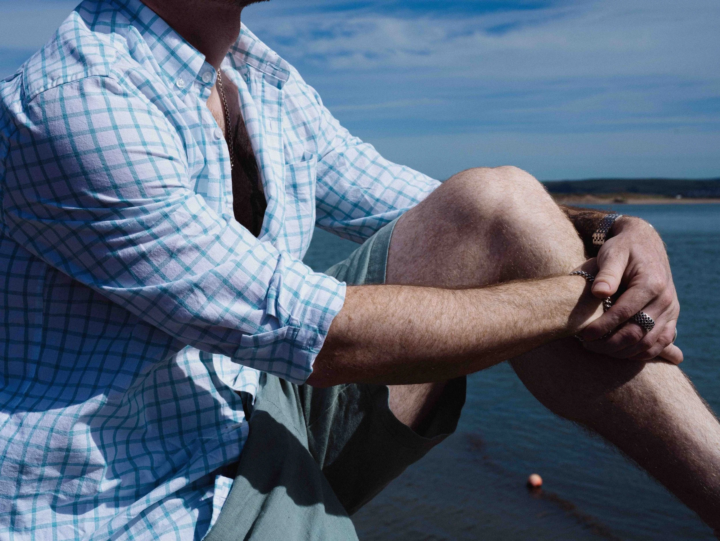A man in a light blue checkered shirt sitting outdoors near water, with his arms crossed over his knees. The sky is partly cloudy.