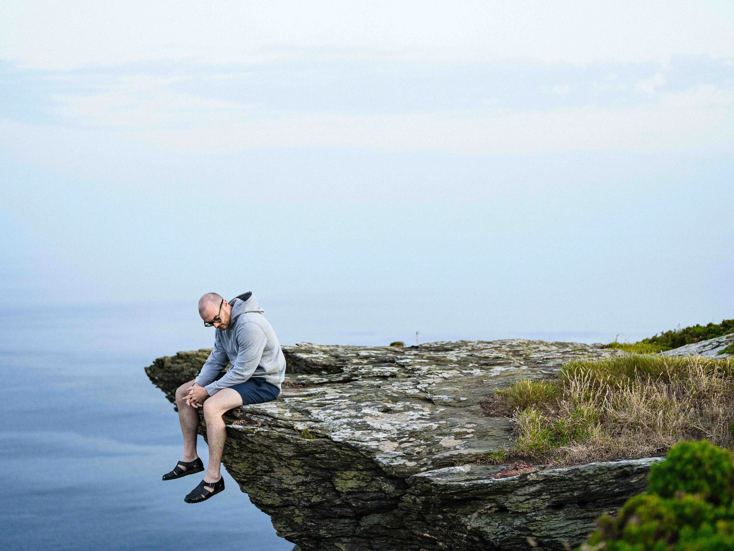 A man sitting on the edge of a rocky cliff by the water, looking down and with his hands clasped, overlooking a body of water under a cloudy sky.