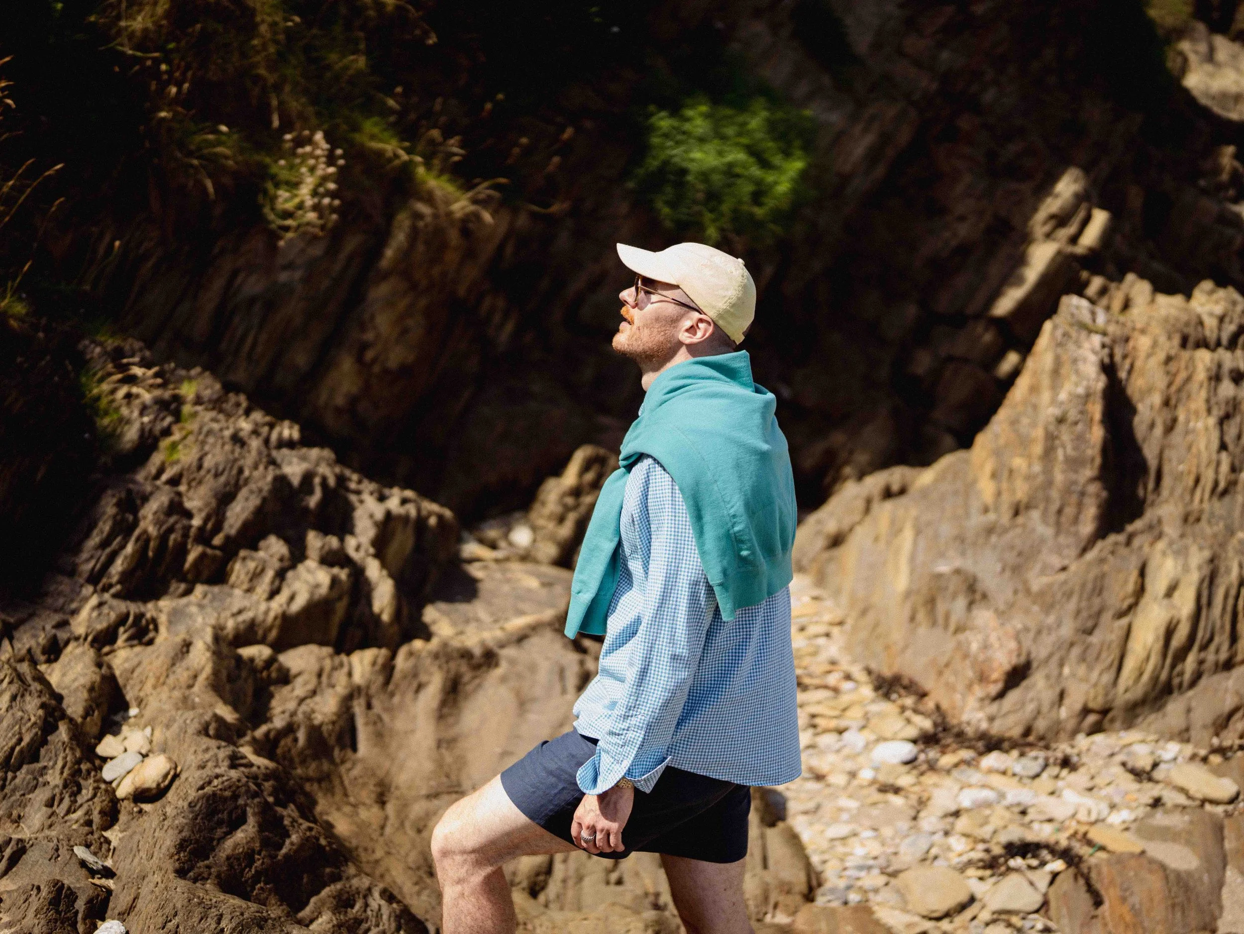 A man with glasses, a beige cap, blue hoodie, and plaid shirt, standing outdoors among rocky terrain with a large rock formation and some green vegetation in the background.