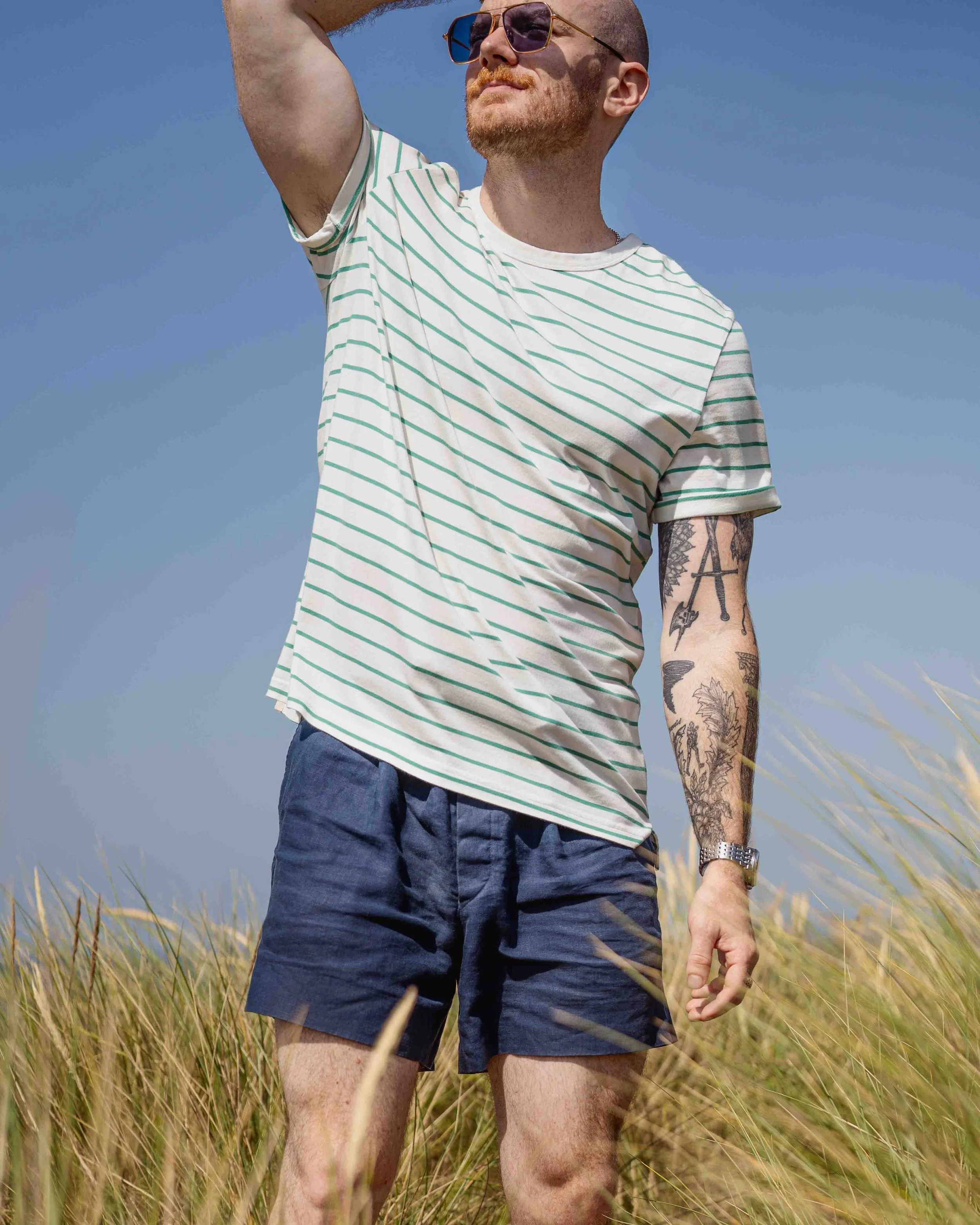 A man with tattoos on his arm wearing sunglasses, a striped t-shirt, and shorts, standing in a field of tall grass under a blue sky.