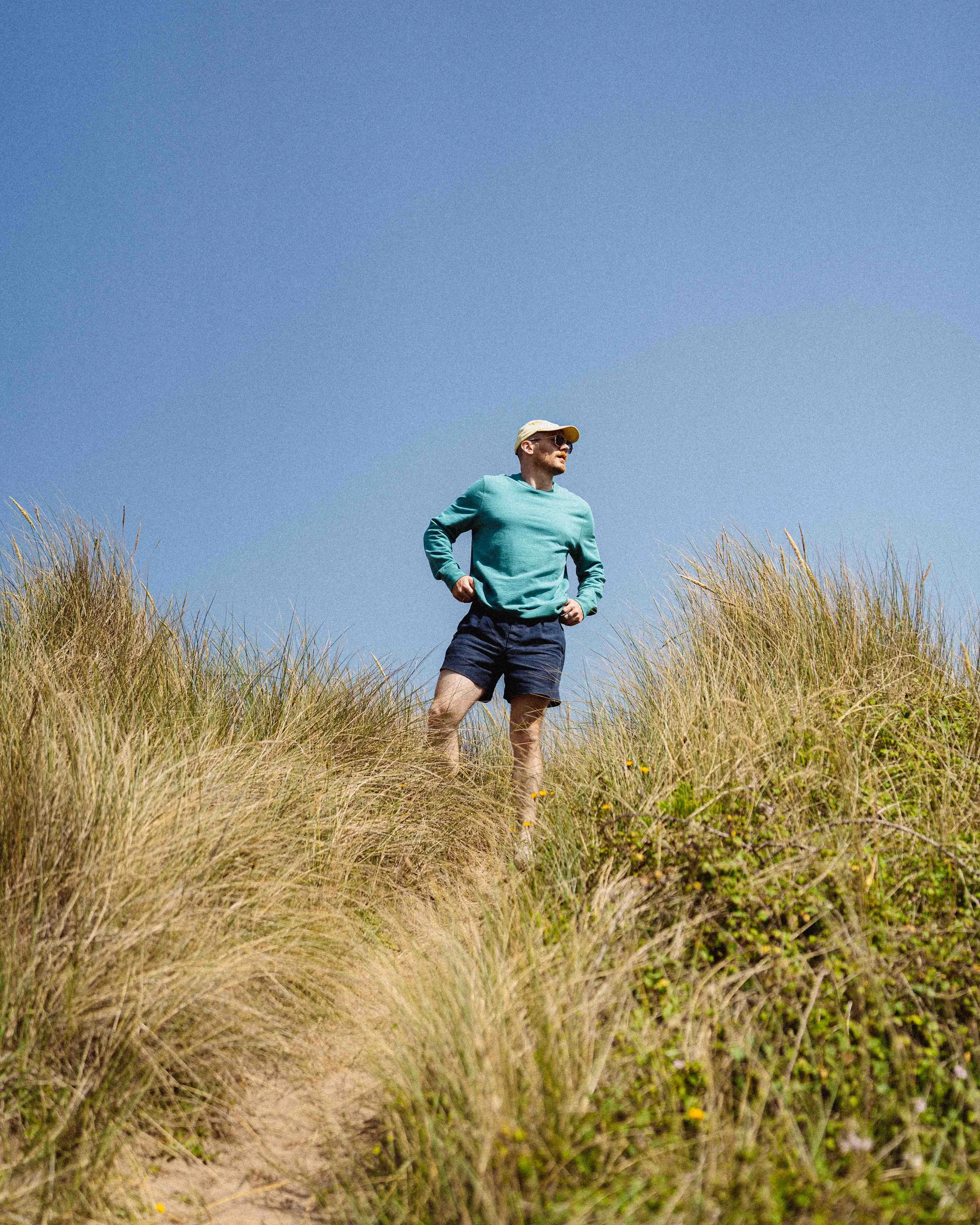 A man wearing a turquoise long sleeve shirt, dark shorts, a beige cap, and sunglasses standing amidst tall, dry grass with a clear blue sky overhead.
