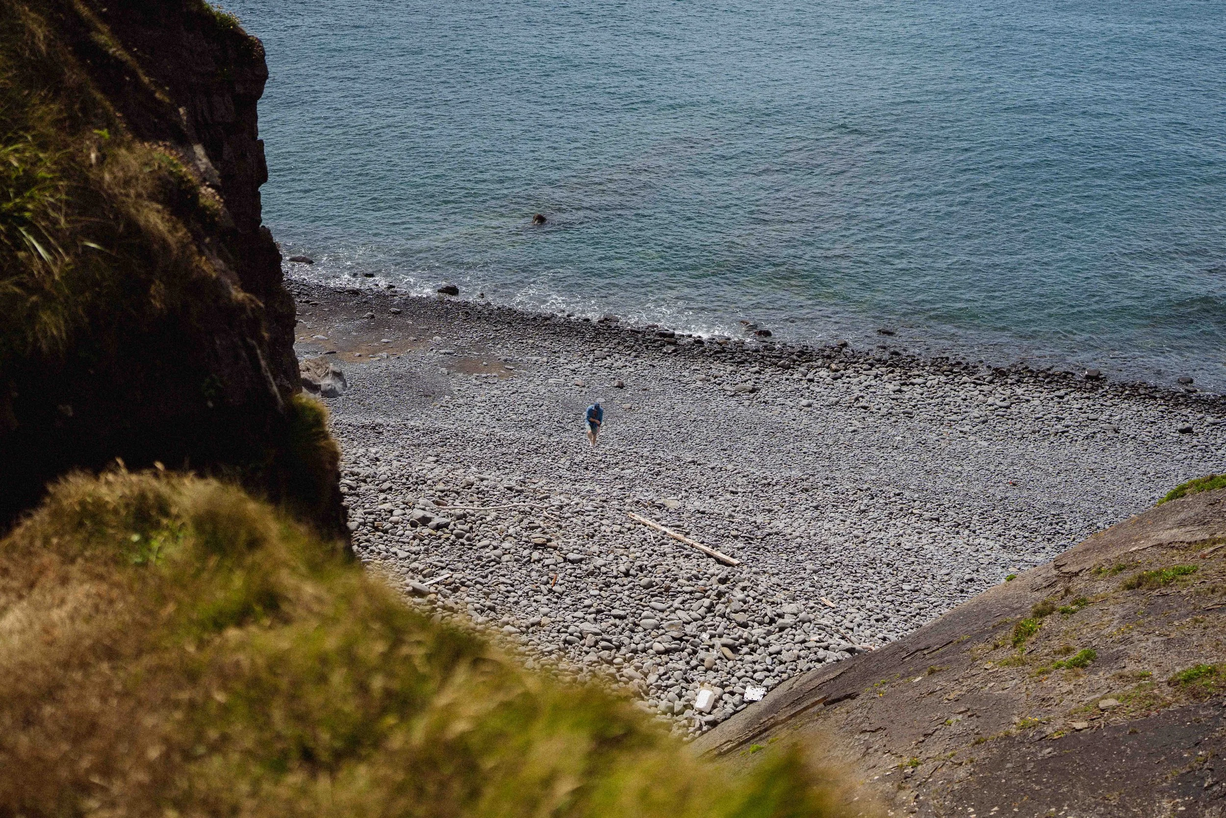 View of a rocky beach with a person walking near the shoreline, seen through a natural frame of moss-covered rocks and grass.