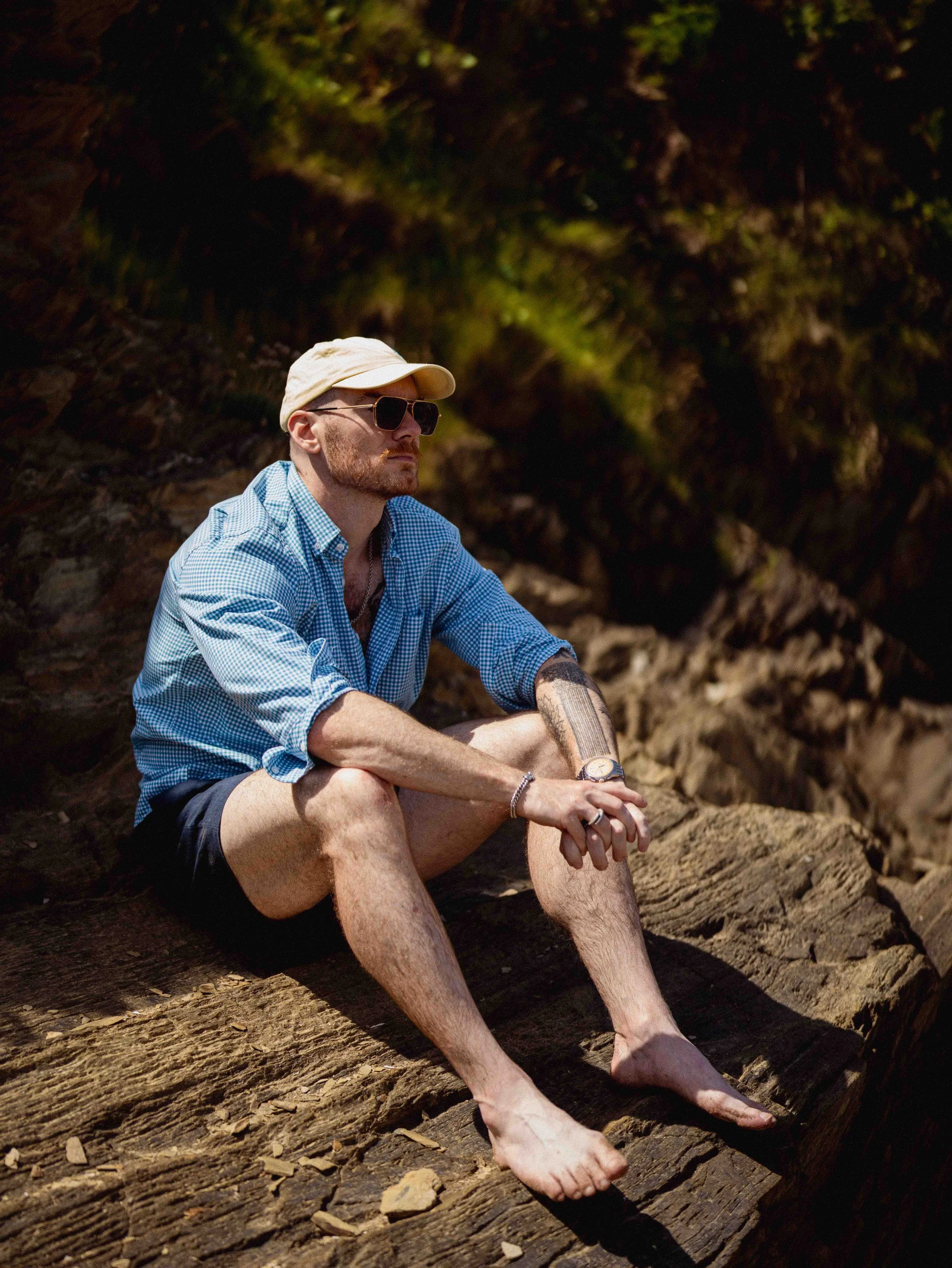 A man with a beard, wearing sunglasses, a beige cap, and a blue checkered shirt, is sitting on a log outdoors during daytime. He has tattoos on his arms and wears a watch and rings on his fingers. He appears relaxed and contemplative against a blurre