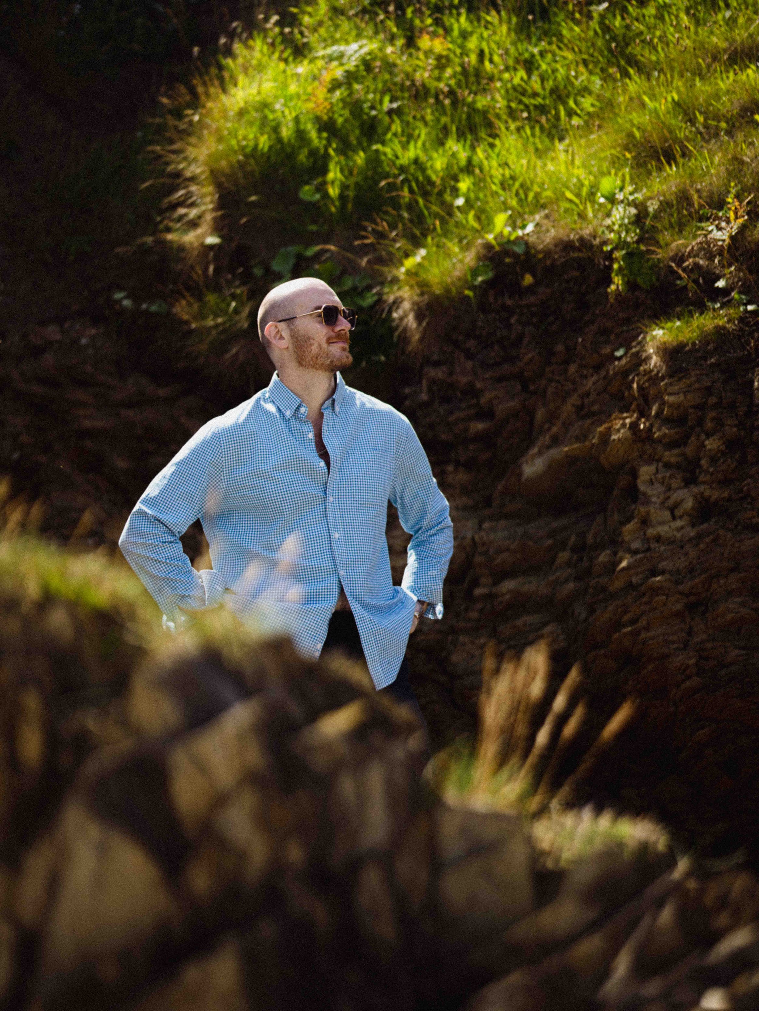 A man with sunglasses and a light blue checkered shirt standing outdoors among plants and greenery.