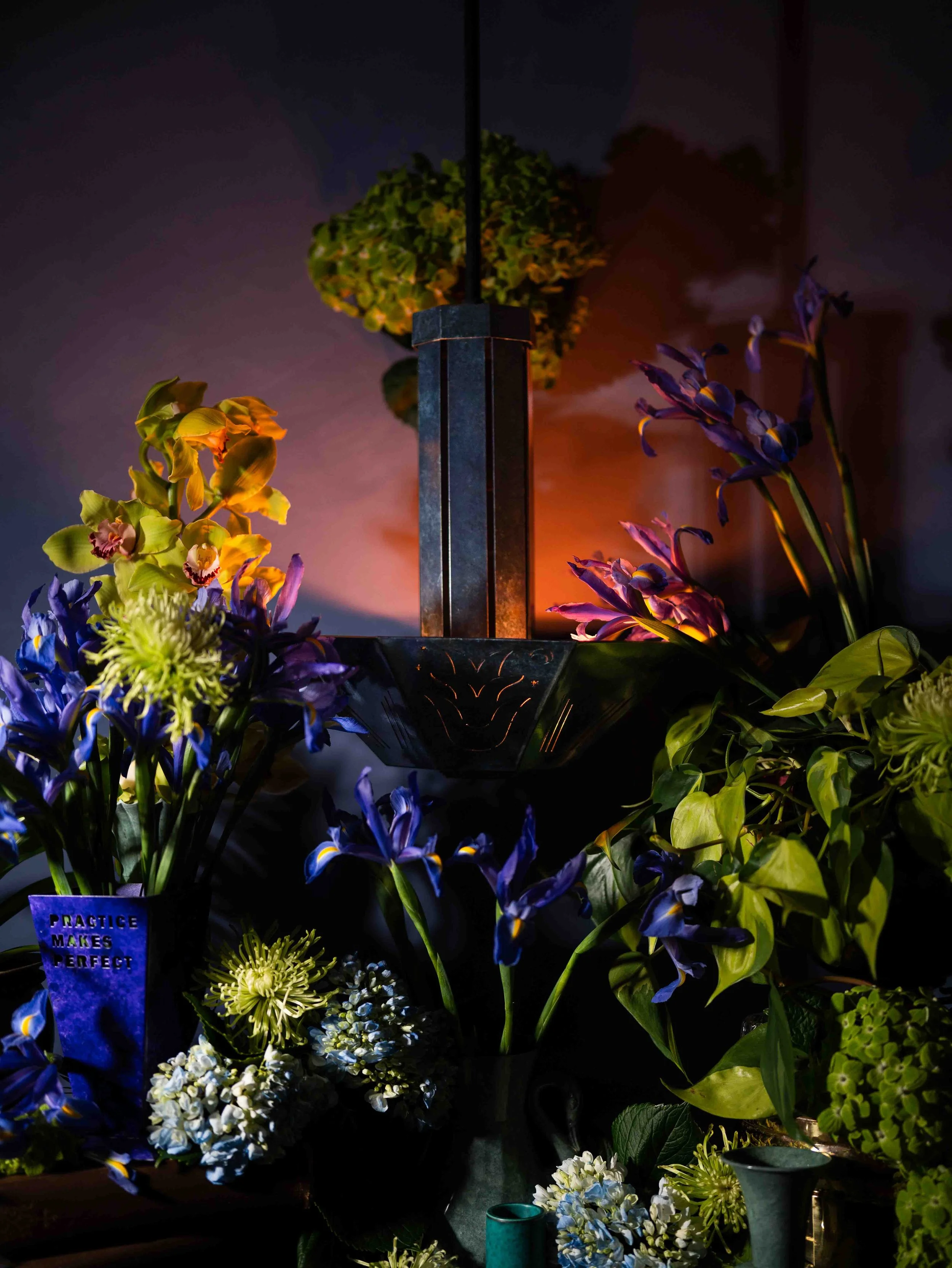 A display of various colorful flowers illuminated by soft lighting, with a sign that reads "Practice Makes Perfect."