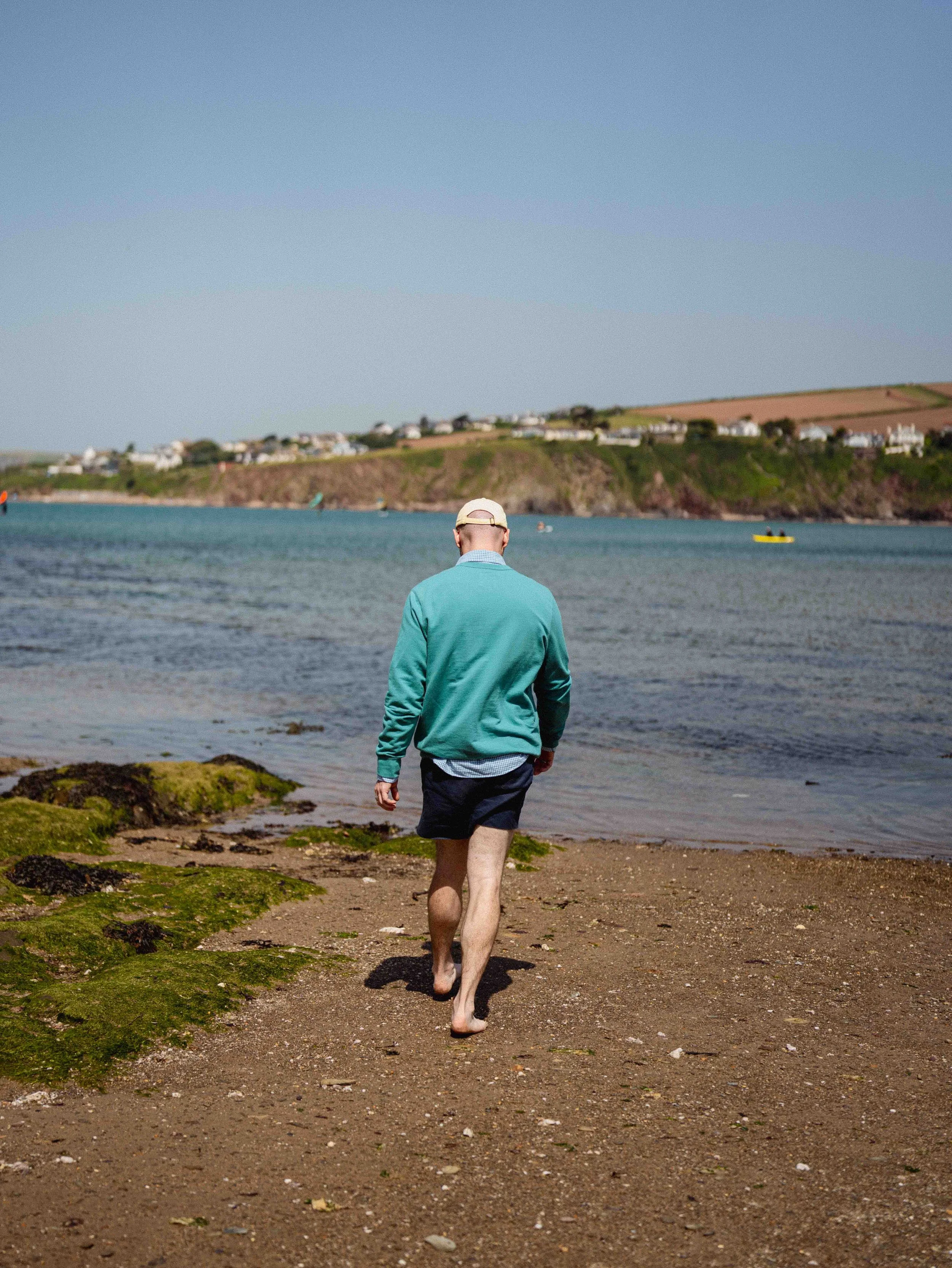 A man walking barefoot along a rocky and sandy shoreline towards the water, with grassy moss patches in the foreground, a bay with boats, and houses on a hillside in the background under a clear blue sky.