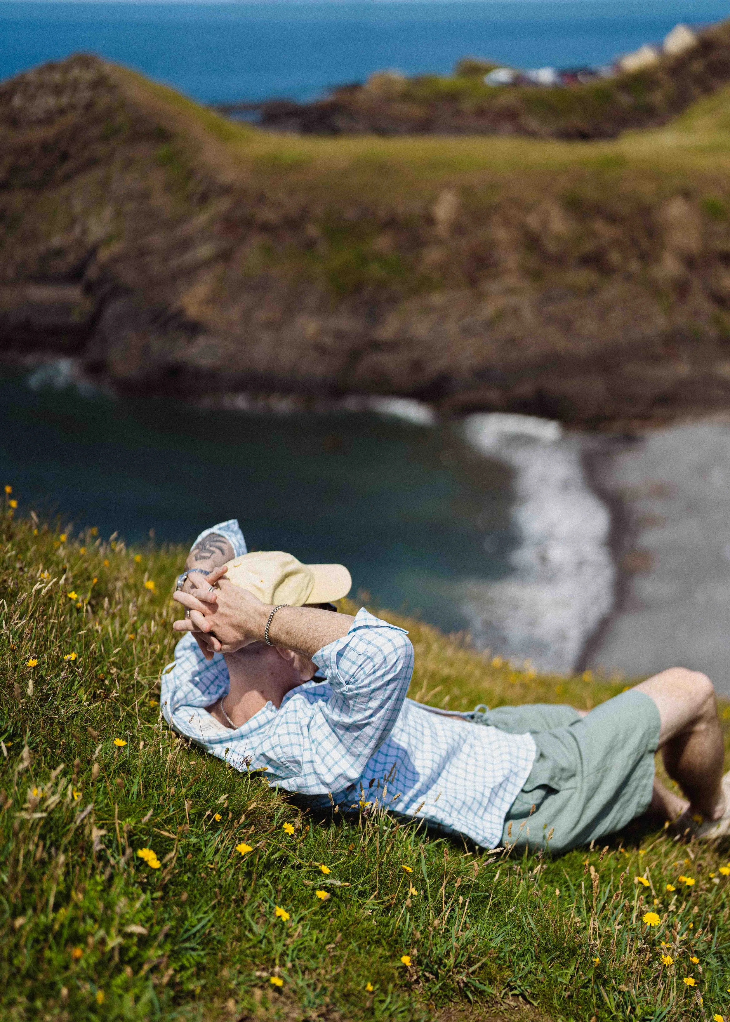 Person lying on grass near coastline with rocky hills in background, wearing a plaid shirt, shorts, beige hat, and holding their hat.