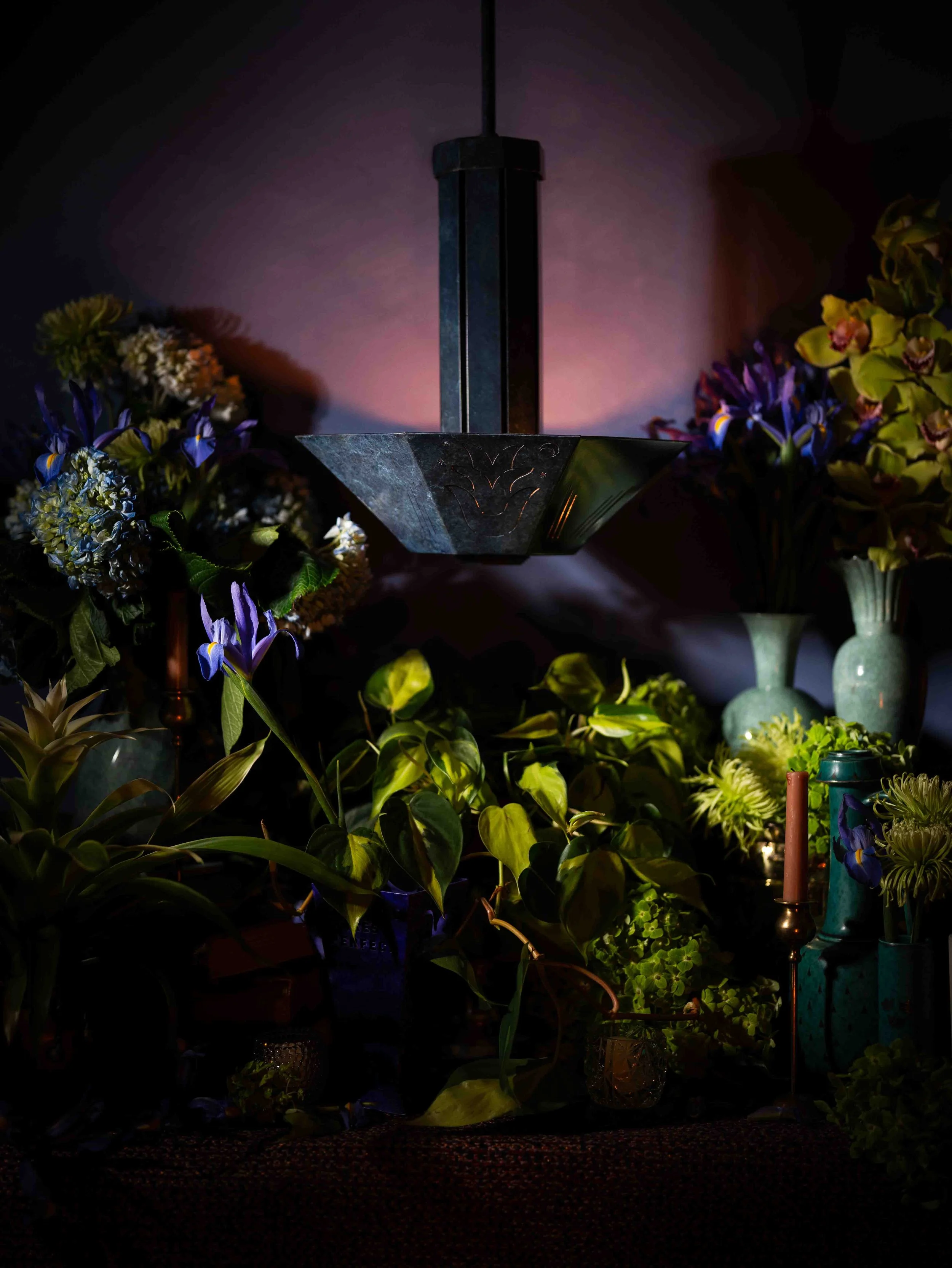 A black geometric hanging lamp surrounded by an arrangement of various colorful flowers and green plants in vases and pots, with soft lighting creating a cozy atmosphere.