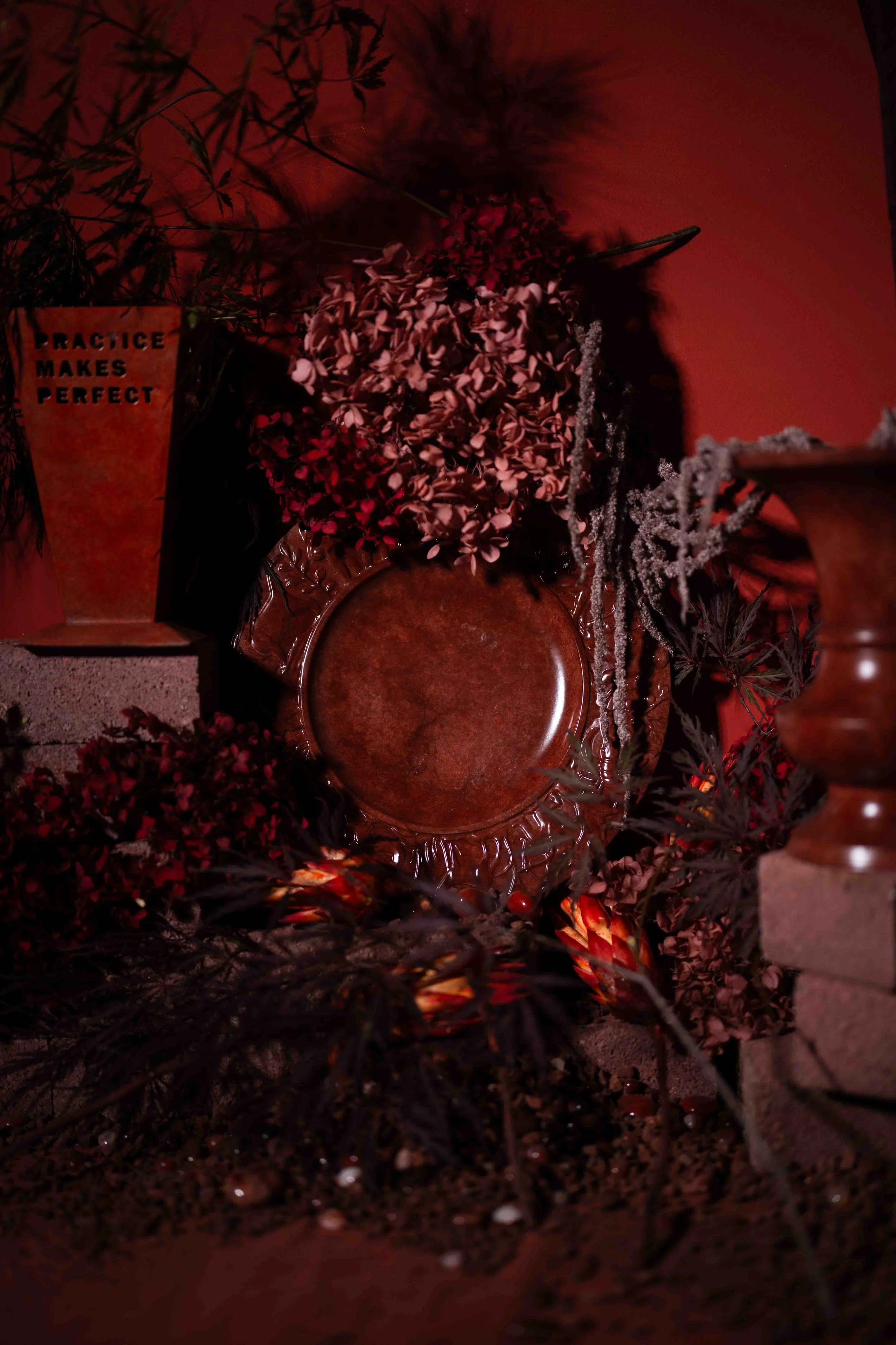 A decorative setting with a sign reading 'Practice Makes Perfect,' arranged with various plants, flowers, and a round wooden tray against a red wall, illuminated in warm lighting.