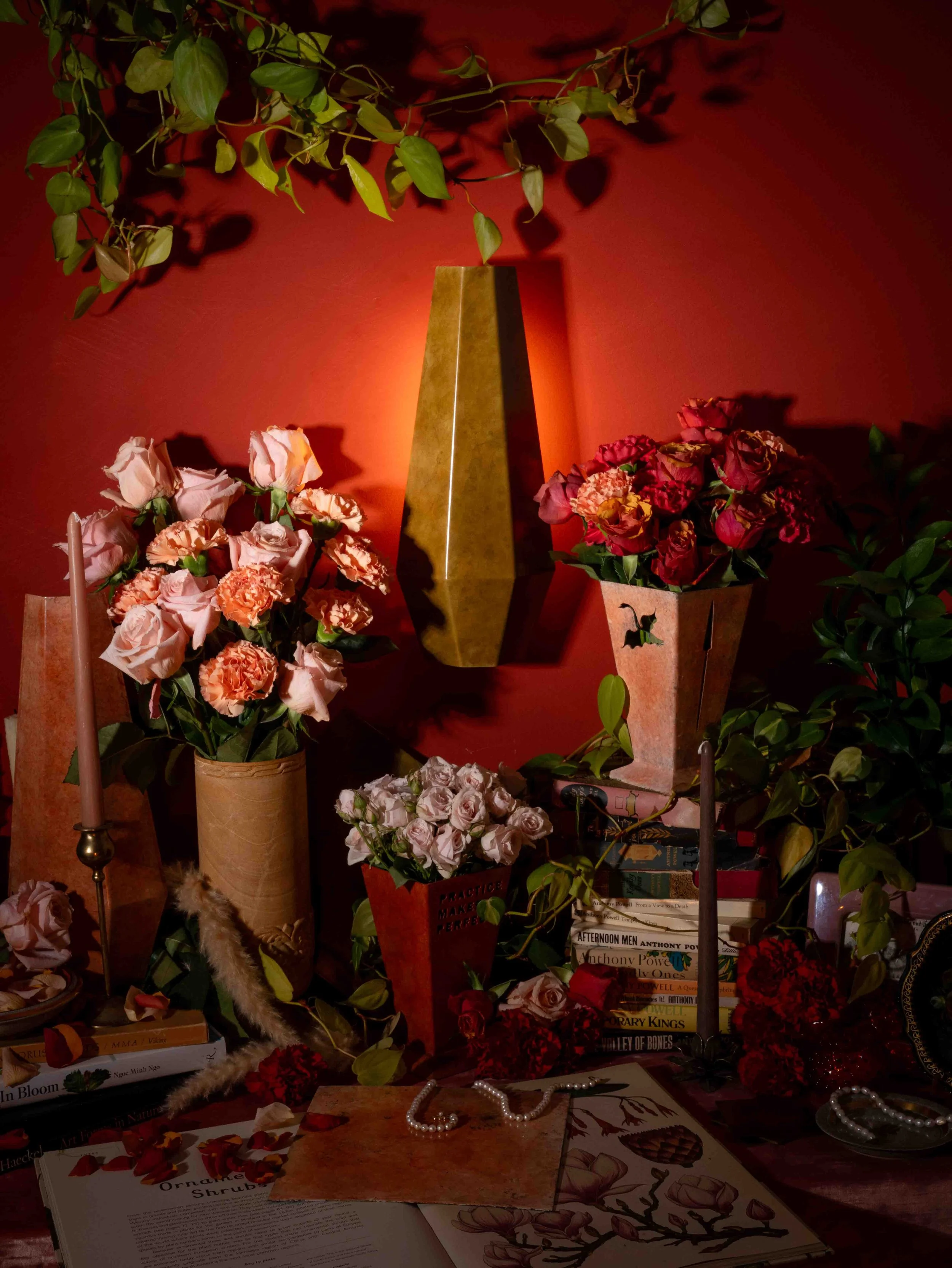 A red wall decorated with a tall geometric gold vase, surrounded by various arrangements of pink and red flowers, green leafy plants, candles, books, and jewelry, creating a warm, cozy, and elegant ambiance.