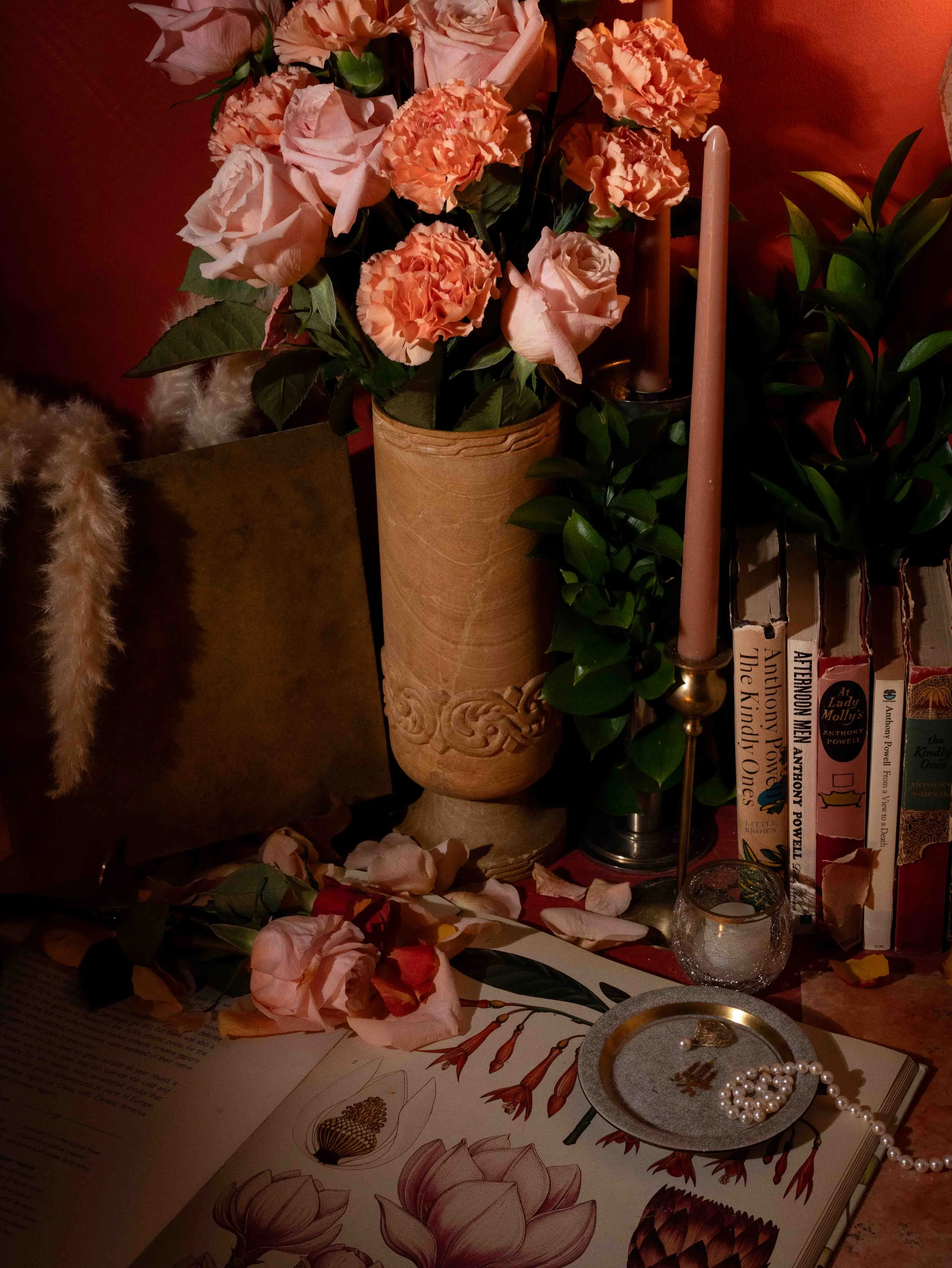 A floral arrangement of pink and peach carnations and roses in a tall, carved terracotta vase. To the right, a pink taper candle in a holder, and to the far right, a green leafy plant. Below, on an open book, there are pink and ivory rose petals, a s