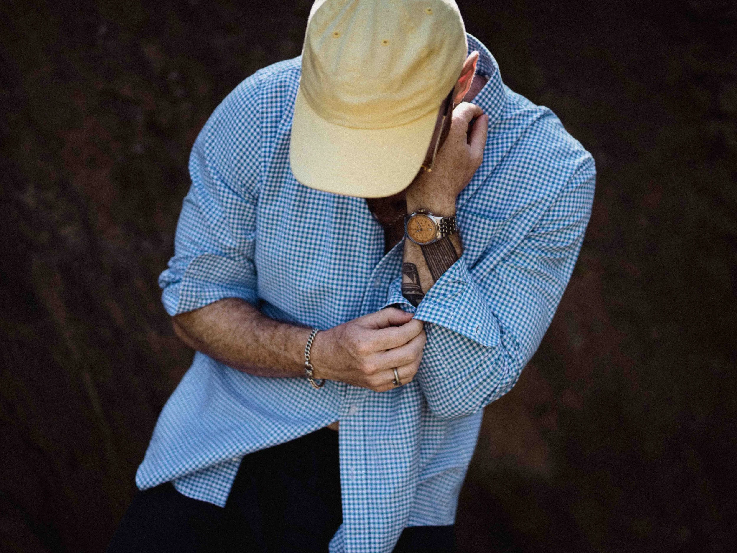 A person wearing a beige baseball cap, glasses, a blue and white checkered shirt, black pants, and a watch, standing outdoors with arms crossed and head bowed.