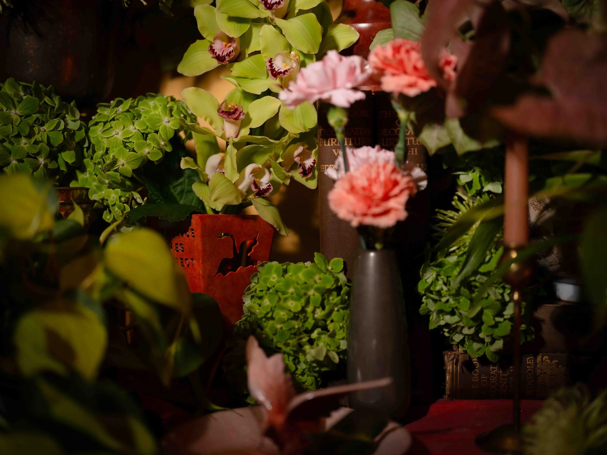 A variety of potted plants and flowers on display, including green foliage, green orchids with striped petals, and pink carnations, arranged in a shop or display setting.