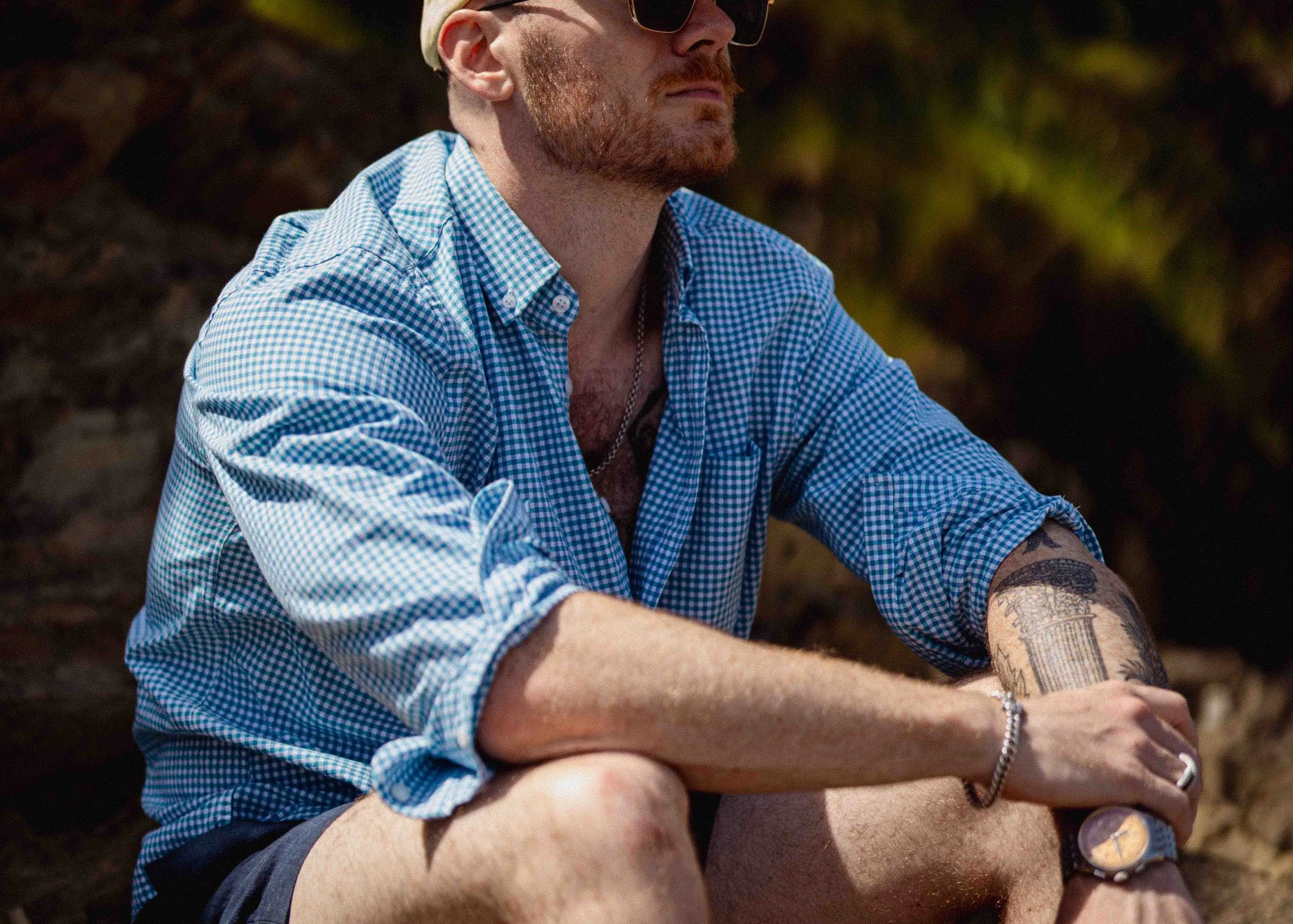 A man with a beard, sunglasses, and tattoos on his arm, wearing a blue checked shirt, sitting outdoors.