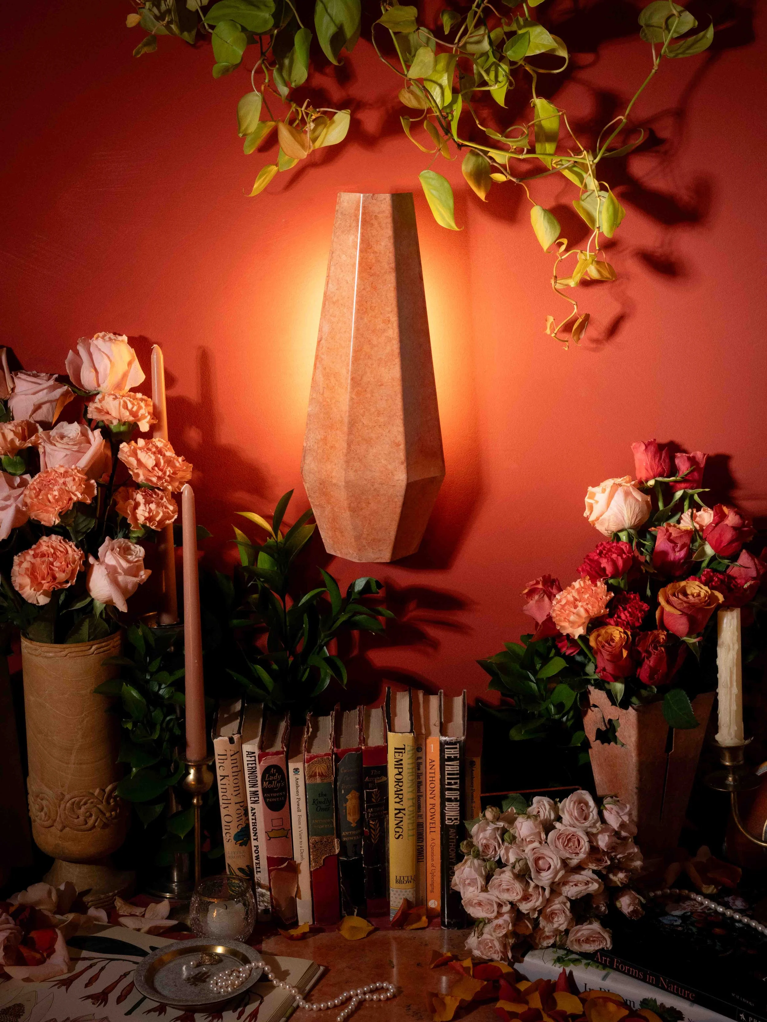 A decorative wall arrangement featuring a tall, oval beige stone vase, numerous pink and red roses, green plants, candles, and a collection of books with floral and nature-themed covers placed on a brown surface, all set against a warm, red-toned wal