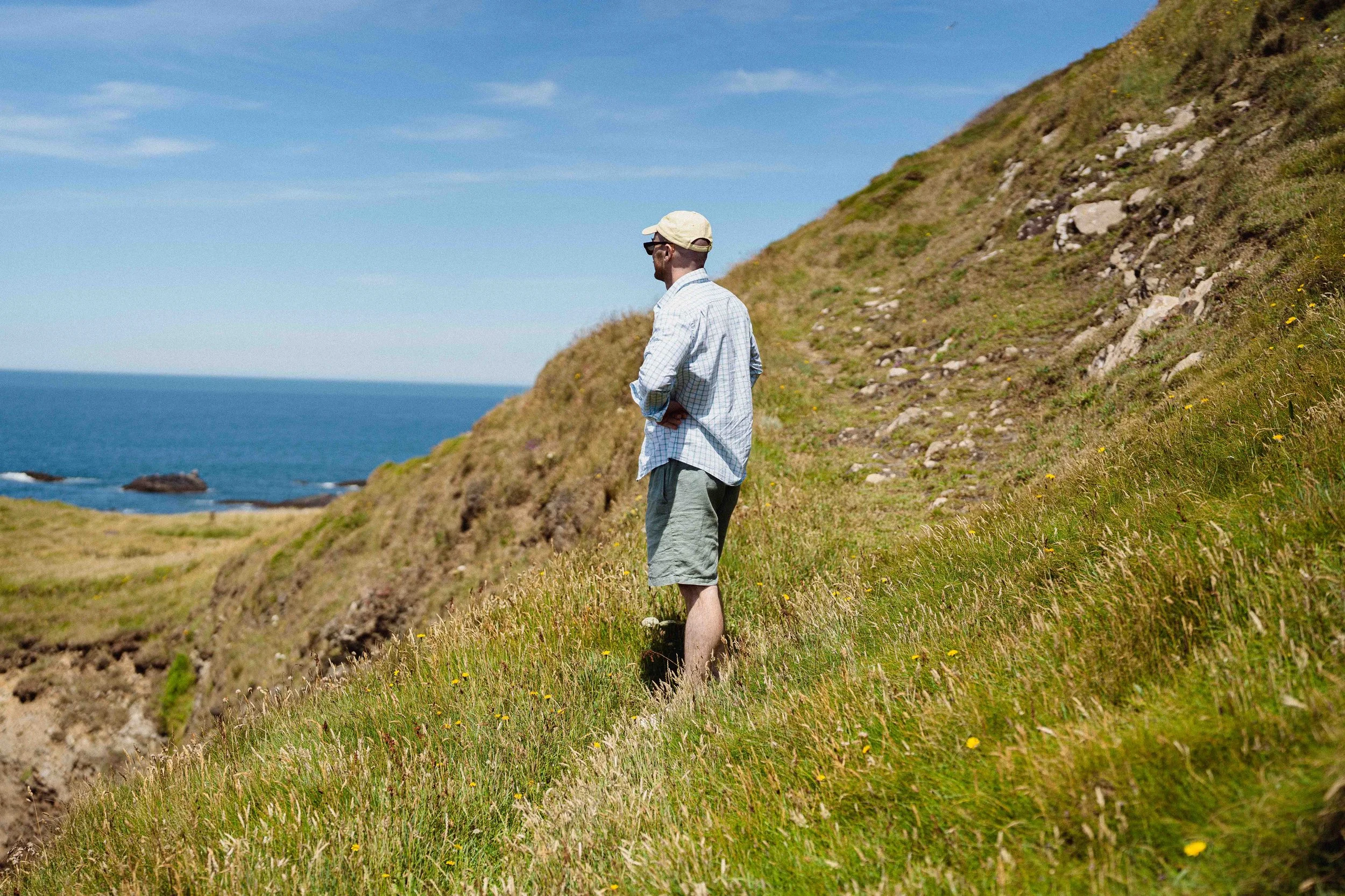 A man in a plaid shirt, shorts, and a cap walking on a grassy hillside near the ocean under a blue sky.